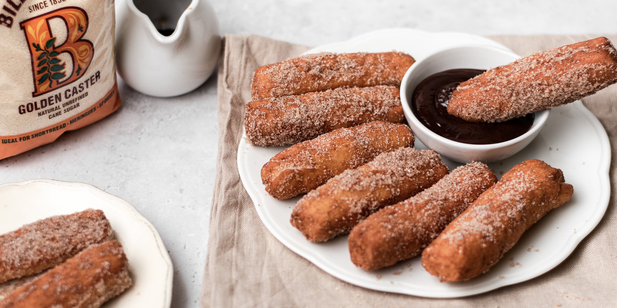 Doughnut Sticks dipping into chocolate dip on a plate, next to a bag of Billington's Golden Caster sugar