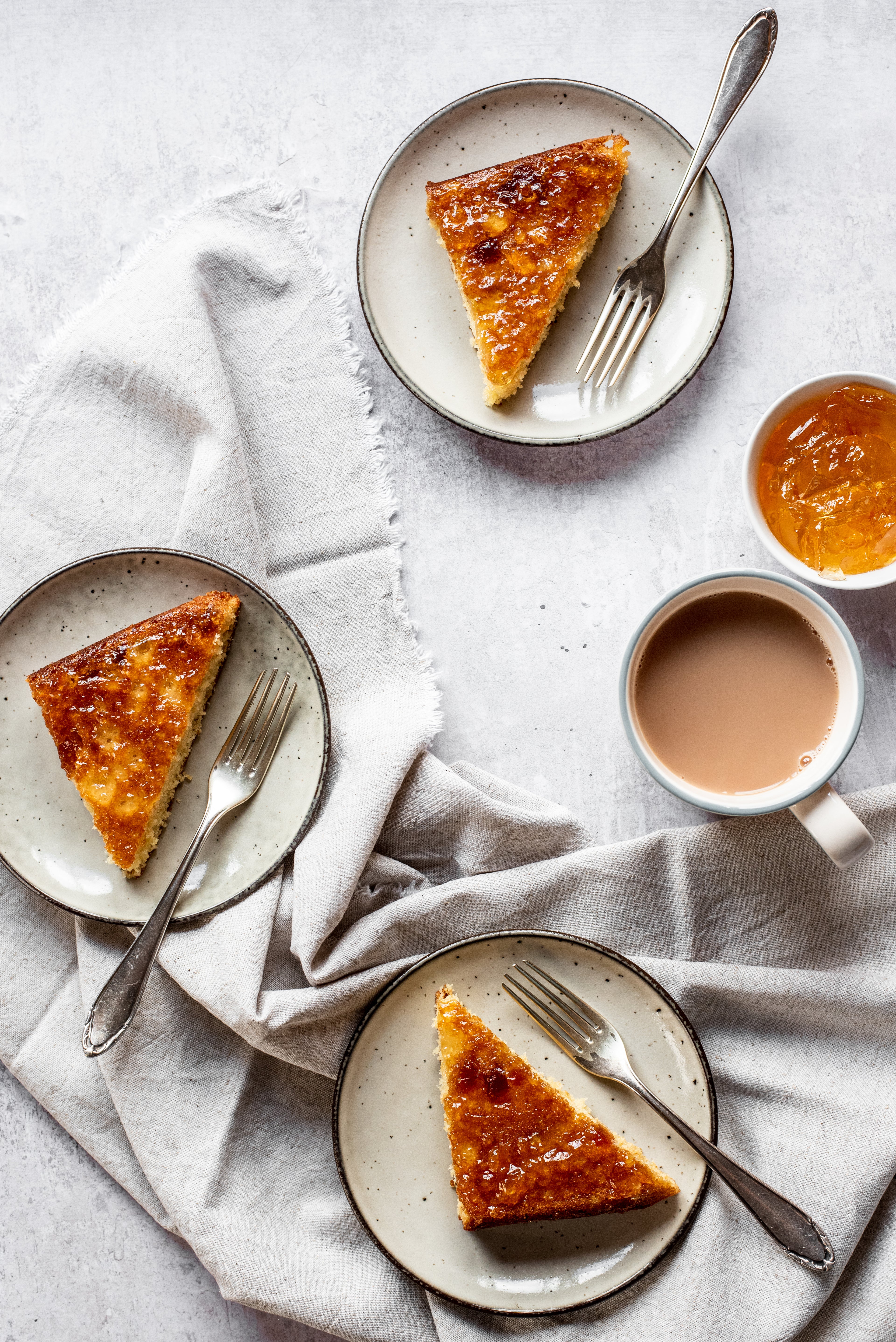 Flay lay of slices of Marmalade Traybake, on plates with forks, on top of linen next to a cup of tea and a bowl of marmalade