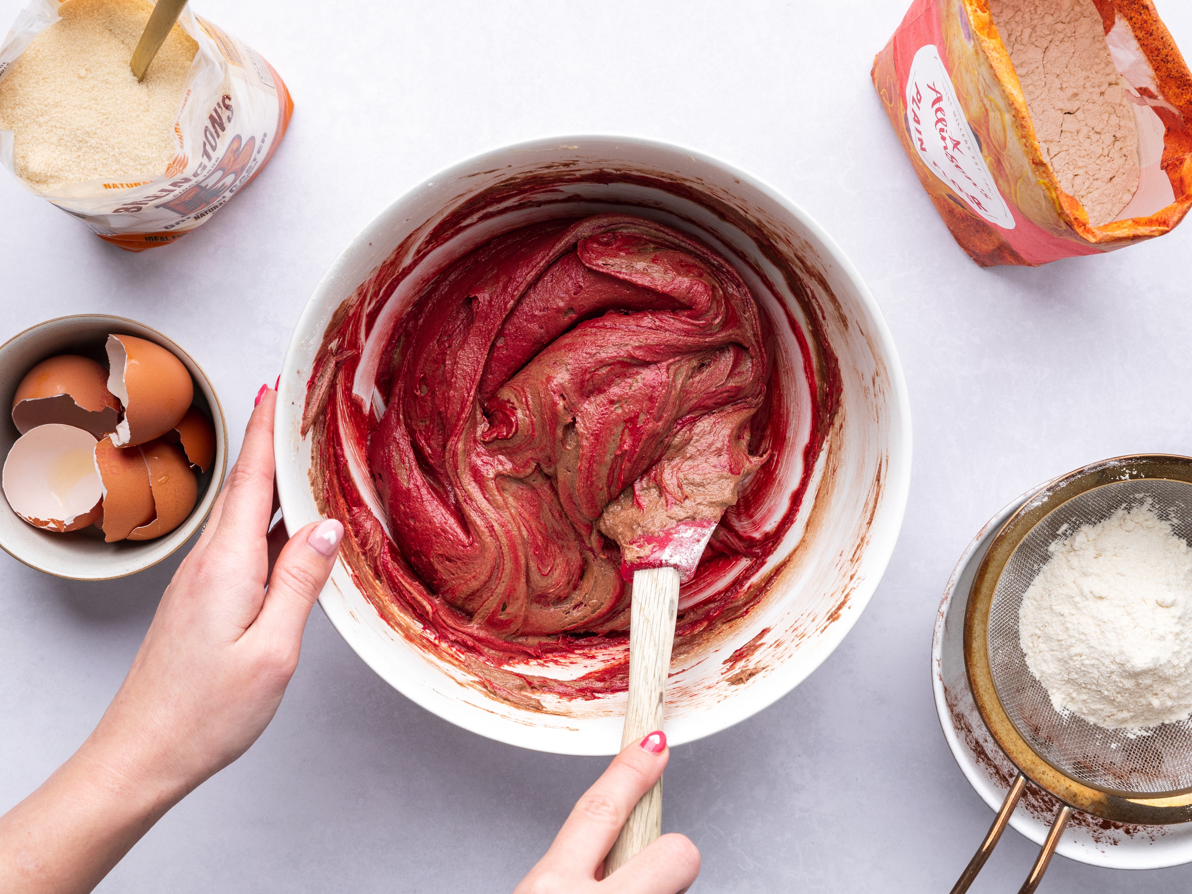 Red velvet cake batter being stirred in a large bowl, beside a sieve of flour and cracked egg shells