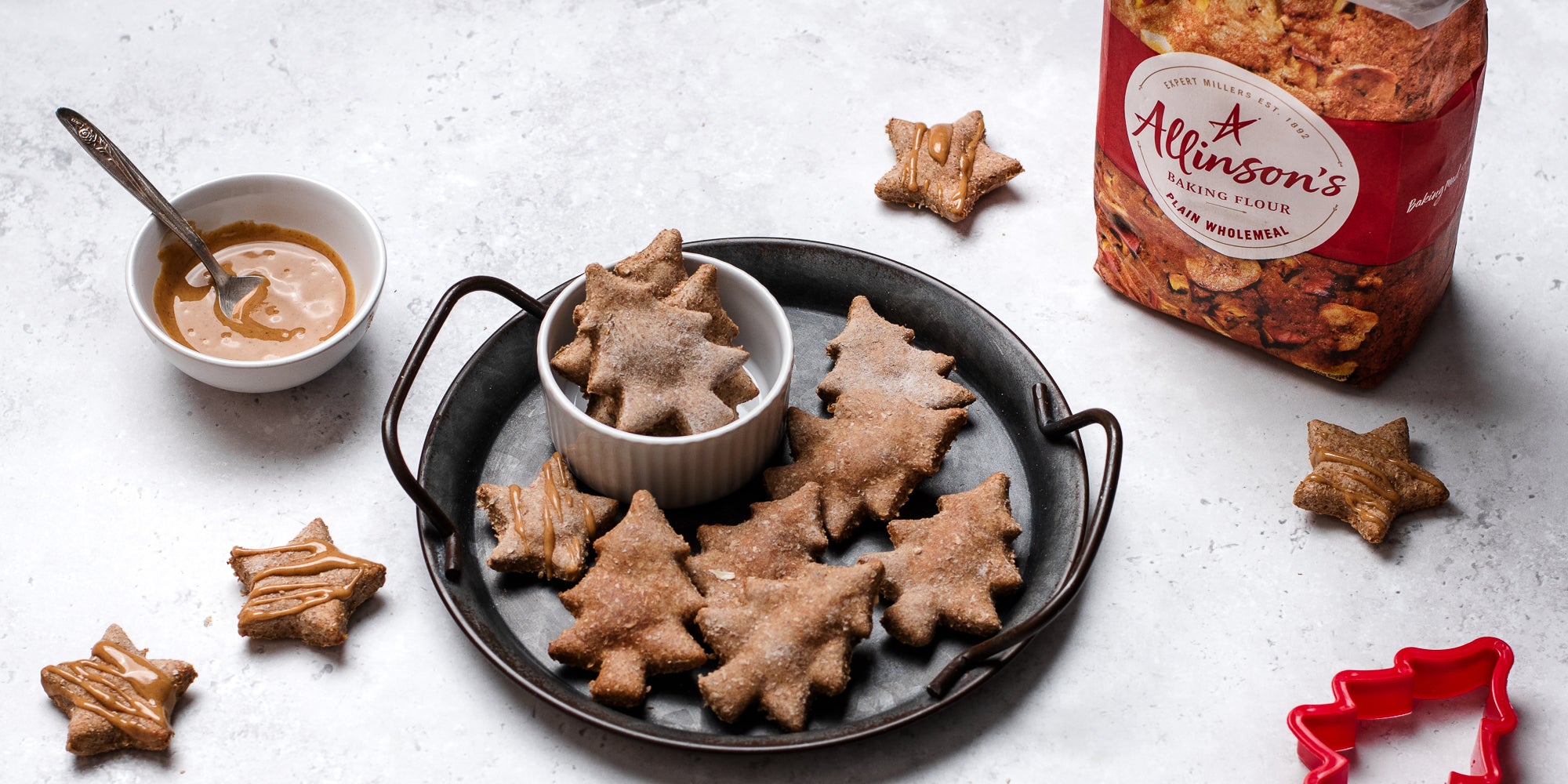 Dog Biscuits cut into festive tree shapes drizzled with Proper Nutty peanut butter next to a bag of Allinsons plain flour