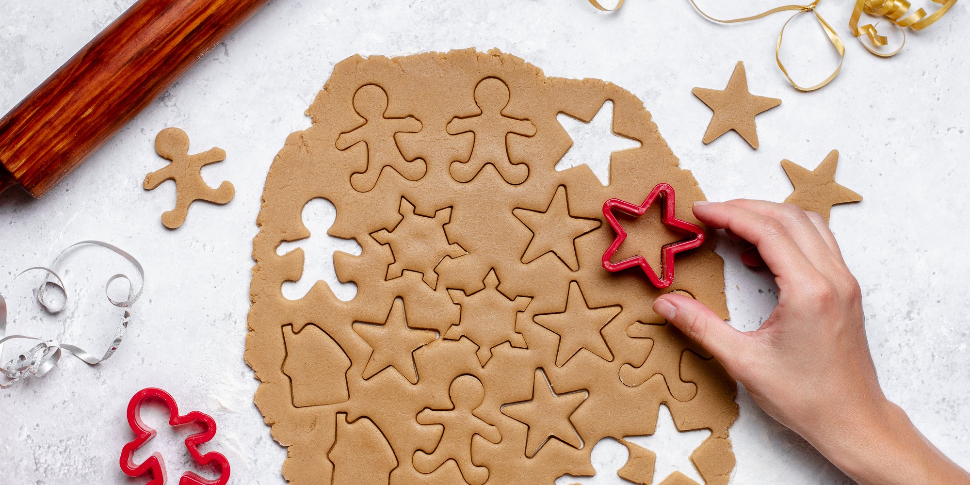 Top view of rolled out Gingerbread Dough with a hand cutting shapes out of it, next to a rolling pin and cut out gingerbread men and stars