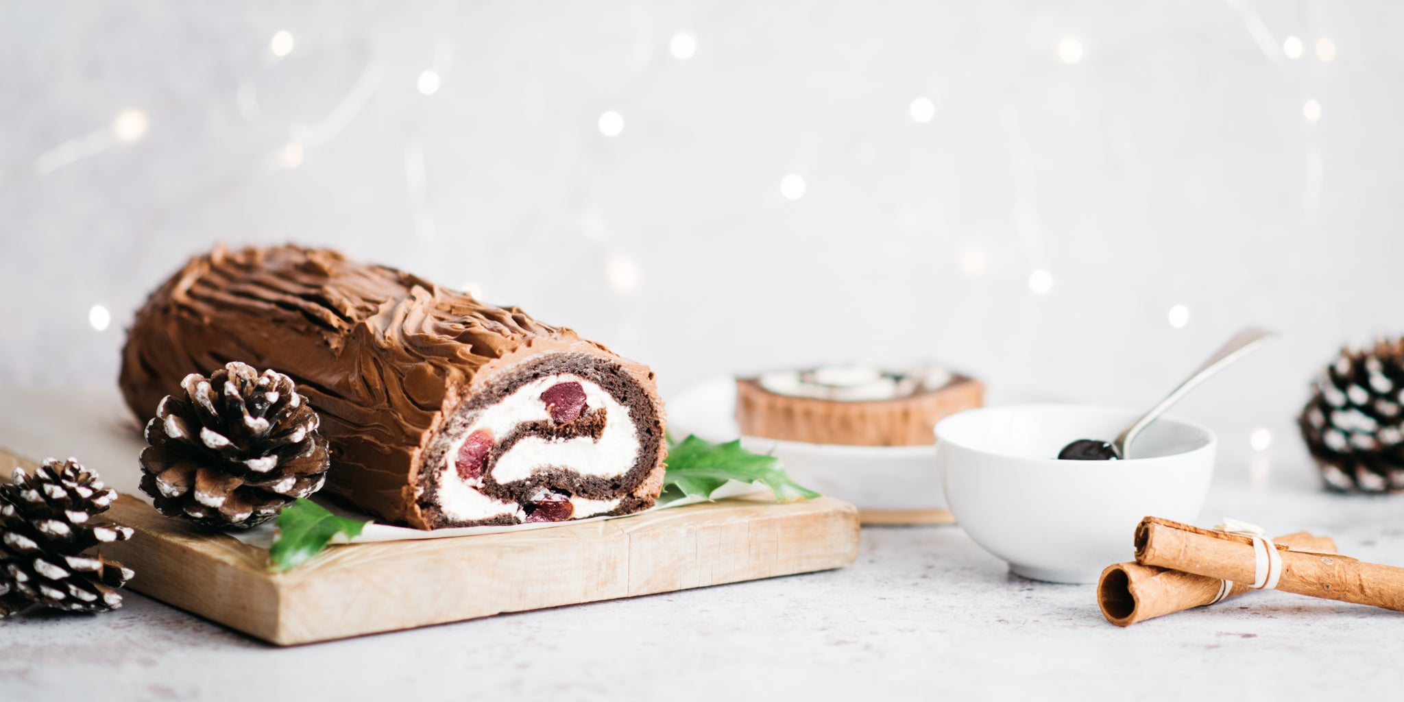 Black forest yule log on wooden board with ingredients and white bowl surrounding it