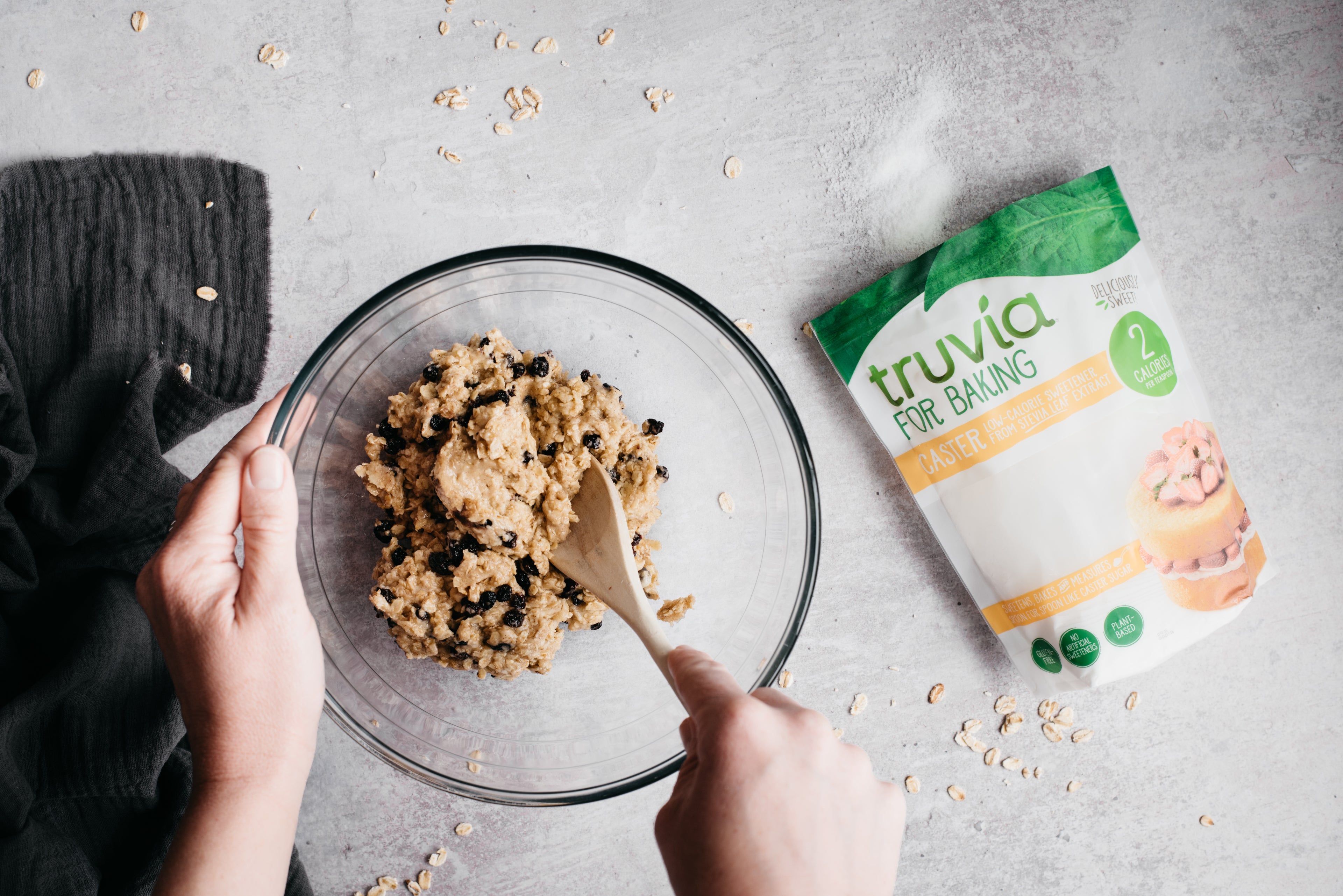Hands mixing Oat & Raisin Cookie dough using a wooden spoon, next to a pack of Truvia for Baking Caster low calorie sweetener