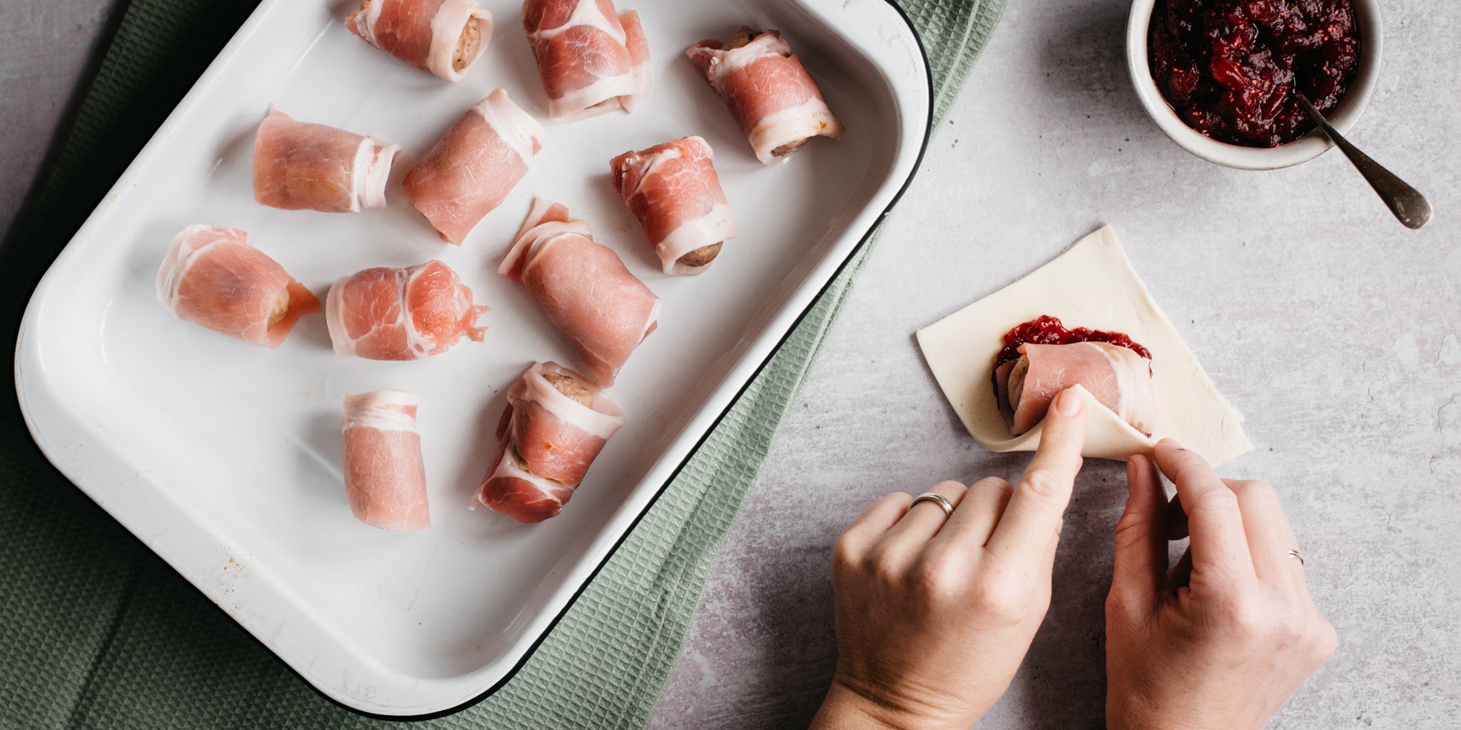 A hand rolling a pig in a duvet next to a tray of wrapped pigs in duvets