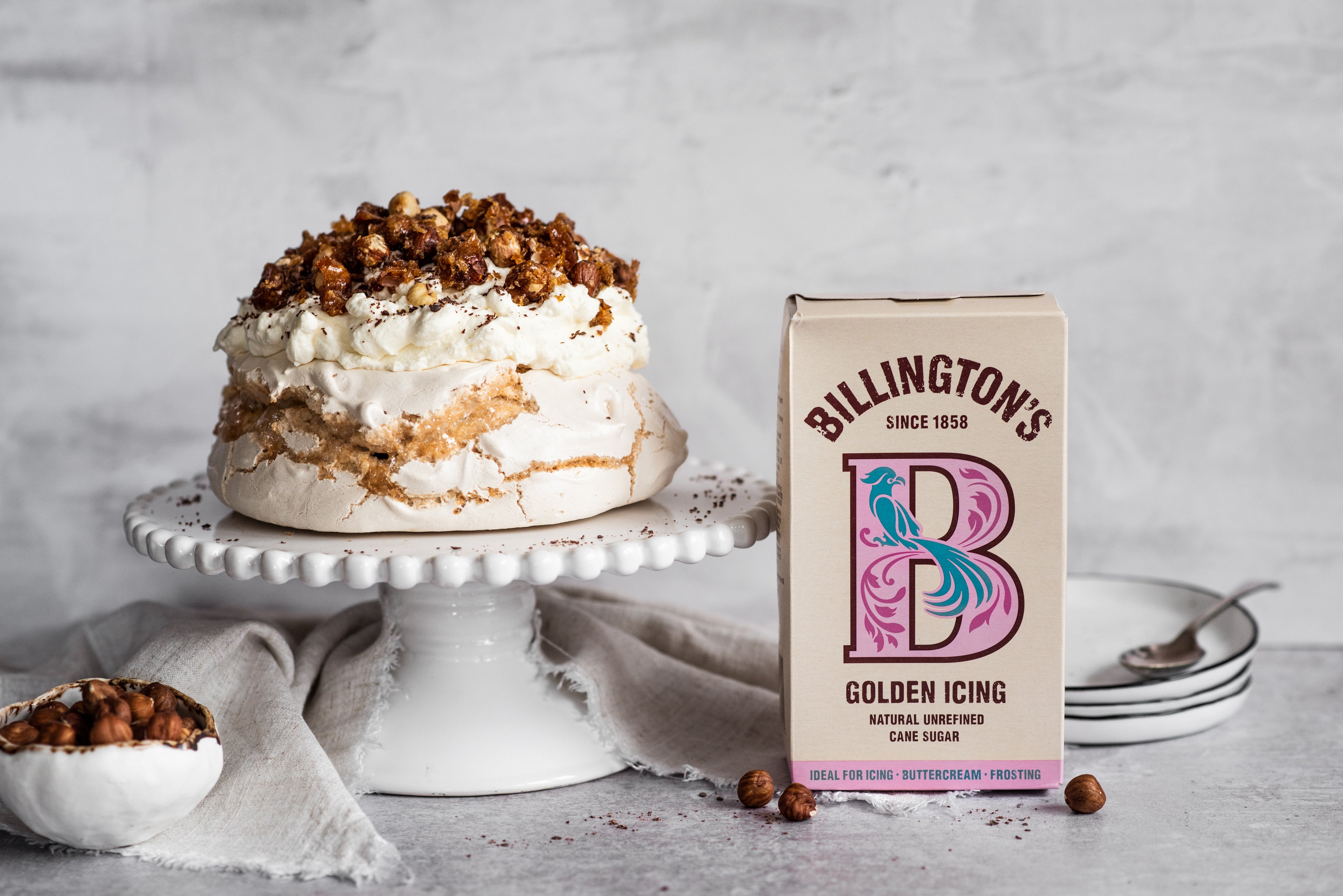 Coffee roasted hazelnut meringue on stand topped with hazelnuts next to icing sugar pack