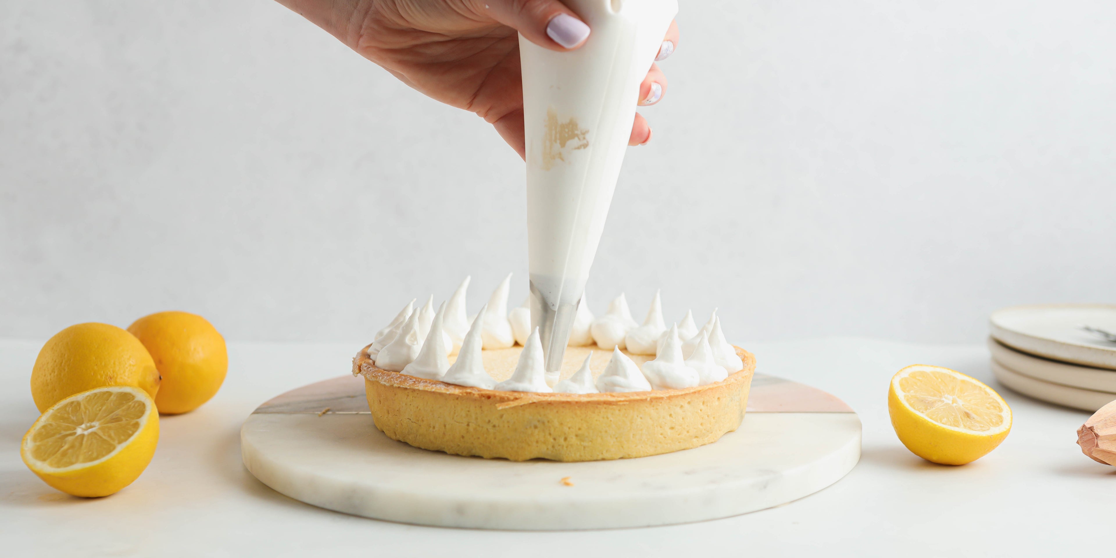 A hand holding a piping bag, piping meringue on top of a lemon pie with lemon slices around it and a stack of whites plates in the background