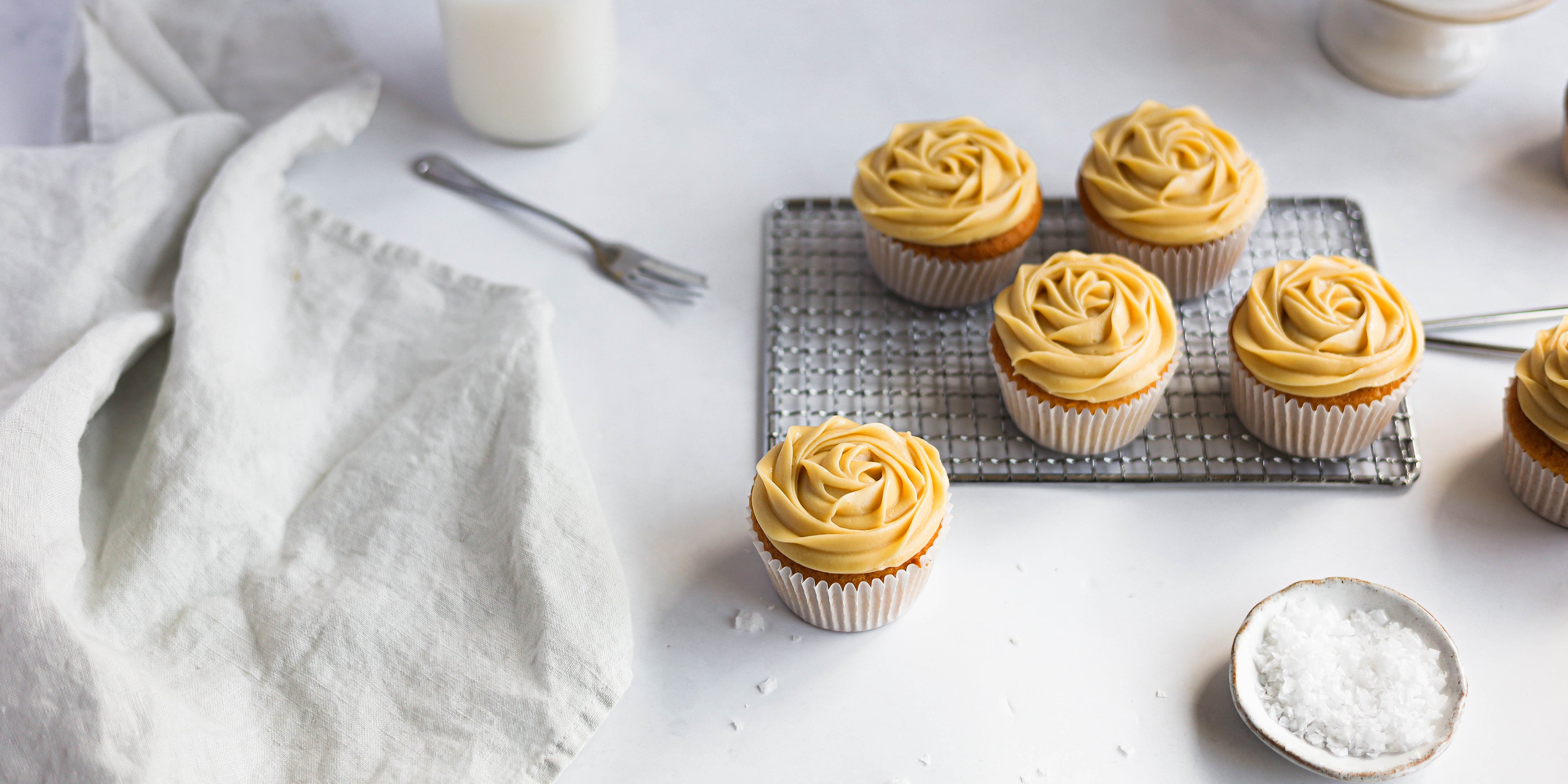 Salted Caramel Cupcake batch on a wire rack with rock salt to garnish and white cloth