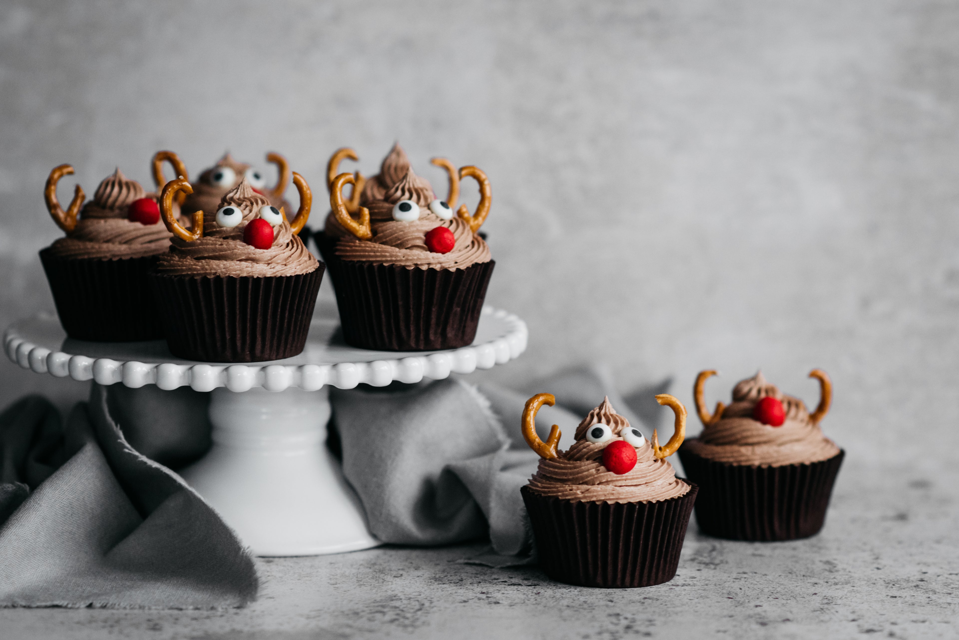 Reindeer Cupcakes on a cake stand, with cupcakes next to it decorated with pretzel antlers and red sweetie noses