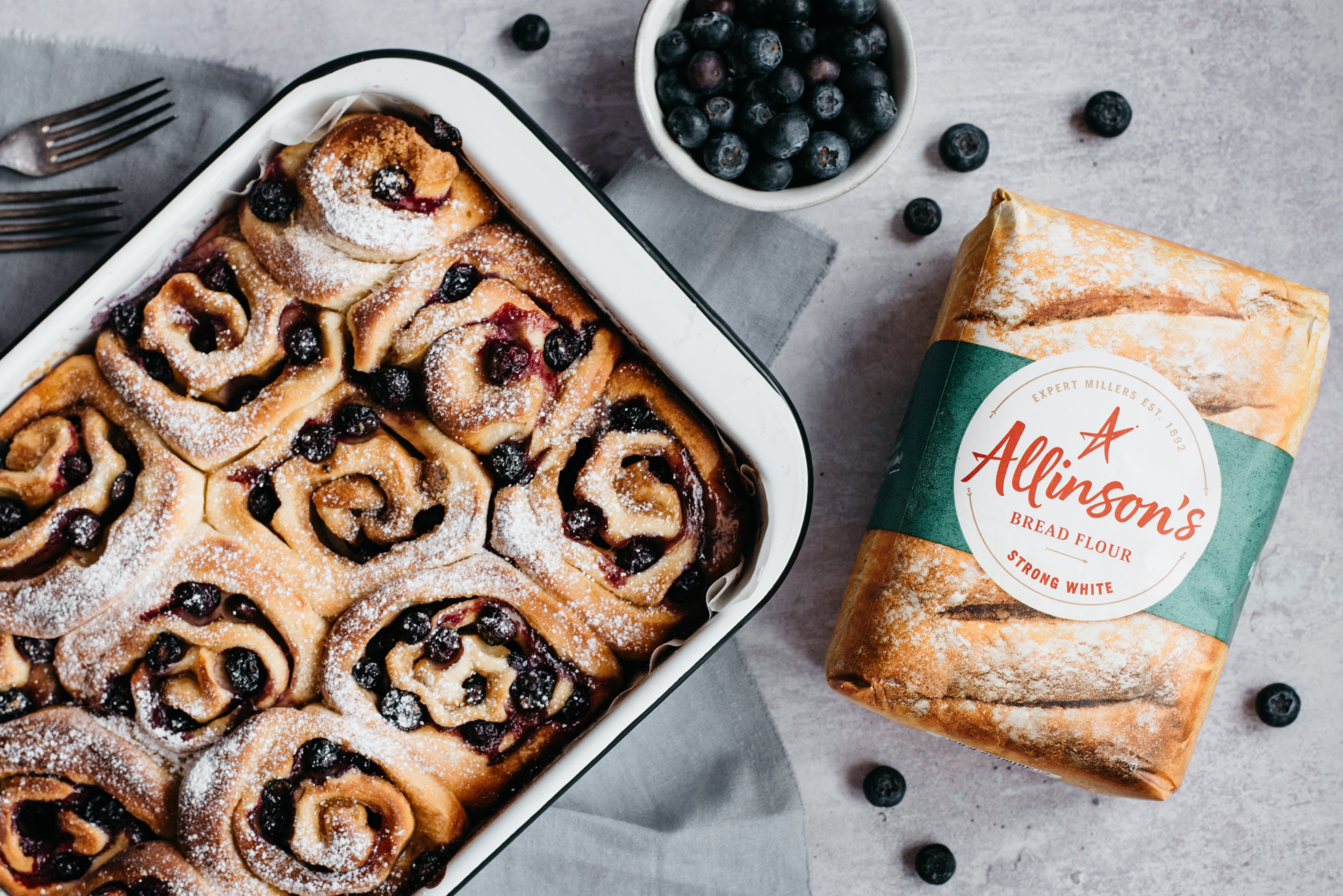 Tray of blueberry and vanilla rolls, dusted with sugar, next to a bag of Allinson's Flour and bowl of blueberries 