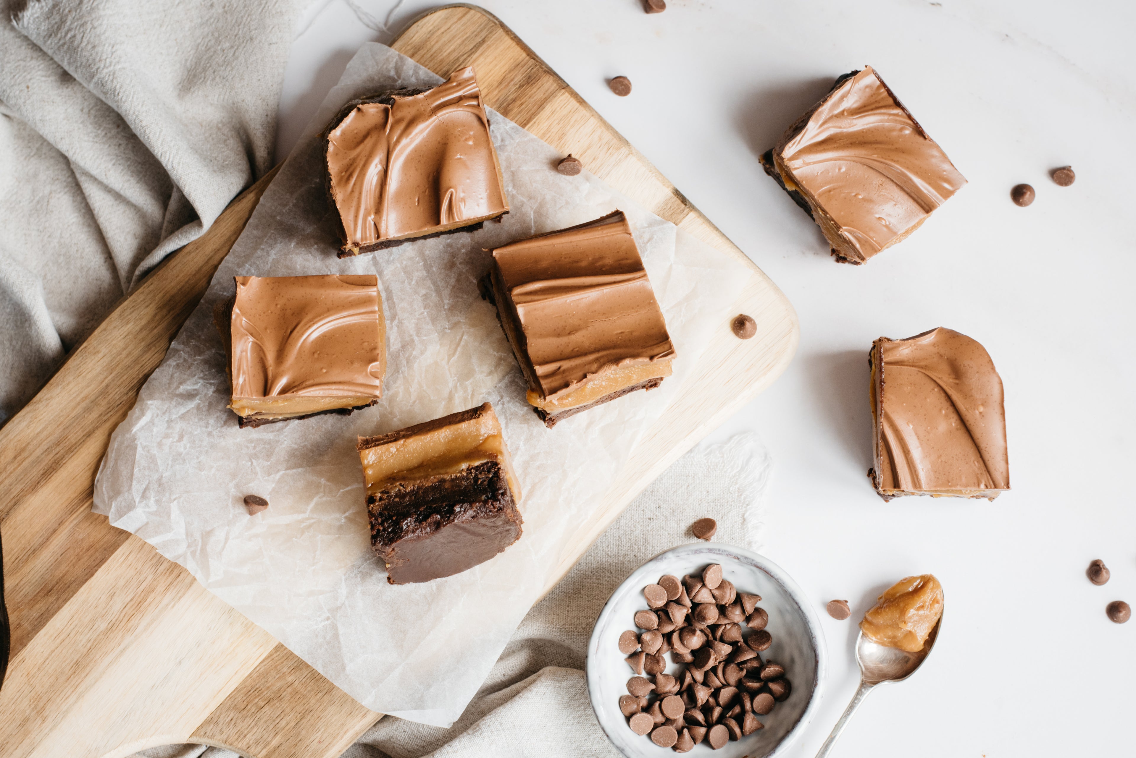 Brownie Based Millionaires Traybake on a wooden serving tray, with baking paper and a bowl of milk chocolate chips