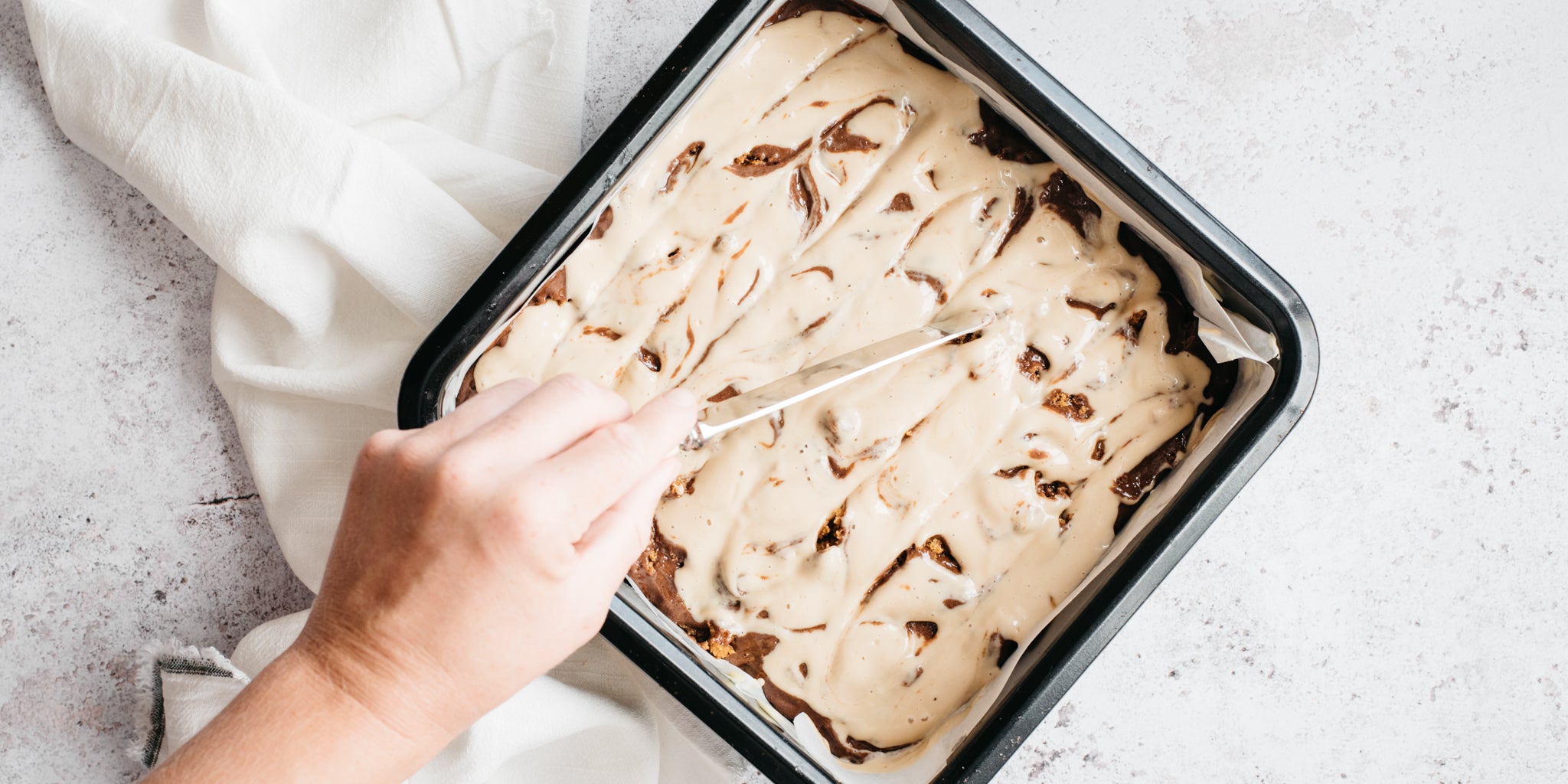 Square tin with brownie mixture and hand stirring the topping in