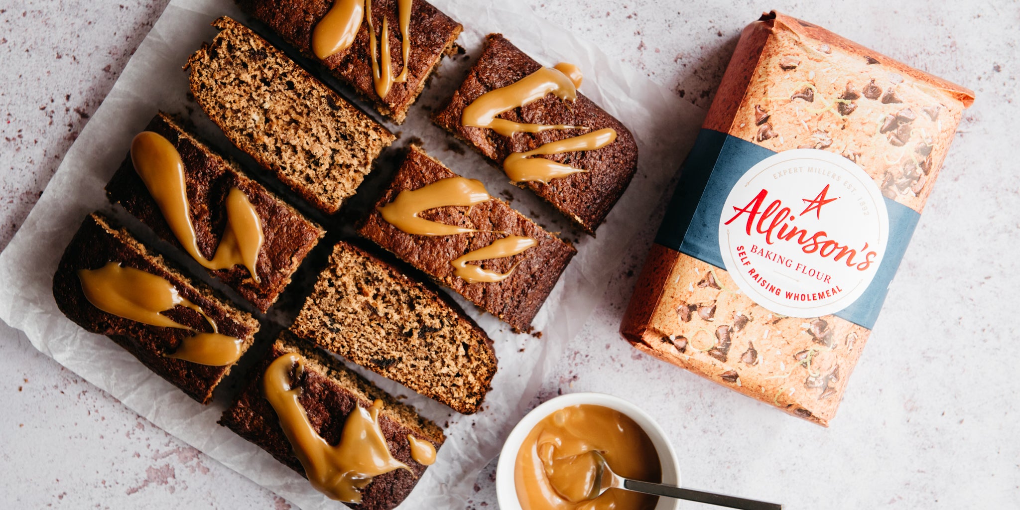 Top view of Ultimate Wholemeal Sticky Toffee Pudding next to a bag of Allinson's wholemeal flour and a dish of toffee sauce with a spoon ready to drizzle