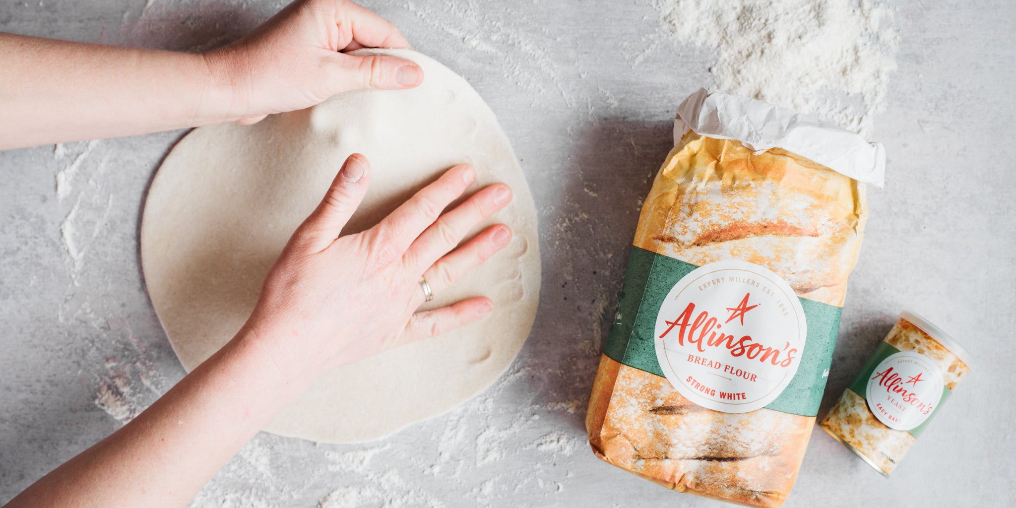 Hands spreading out pizza dough alongside a pack of flour and yeast