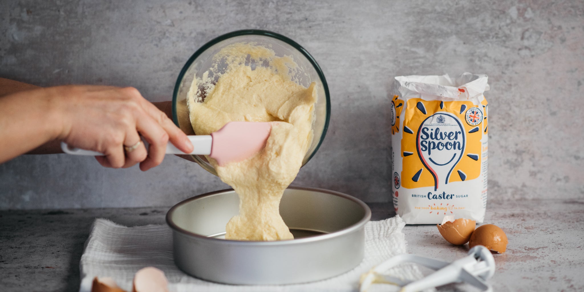 Gluten Free Victoria Sponge batter being poured with hands holding a silicone spatula into a lined baking tray, next to a bag of Silver Spoon Caster Sugar