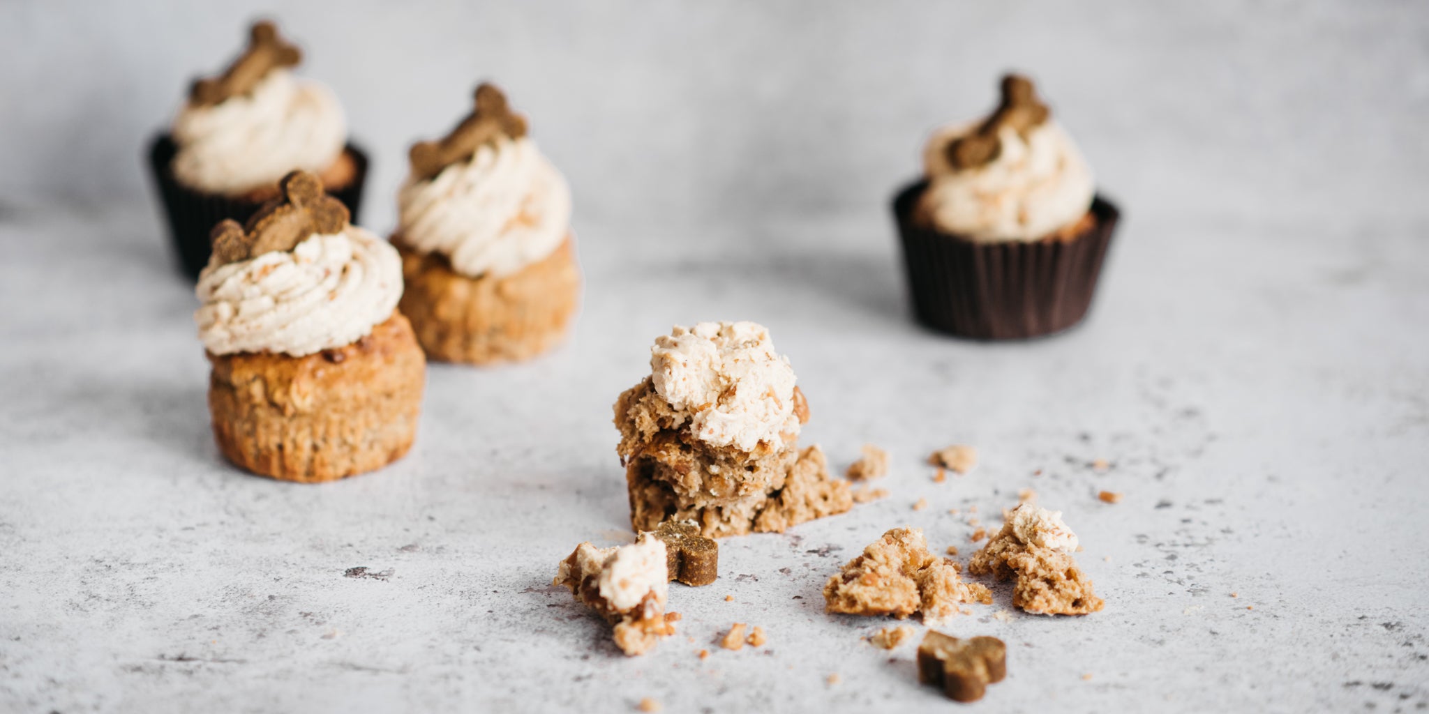 5 cupcakes frosted in shot, two in brown paper cases and two without. One cupcake crumbled in the foreground as though bite has been taken out of it