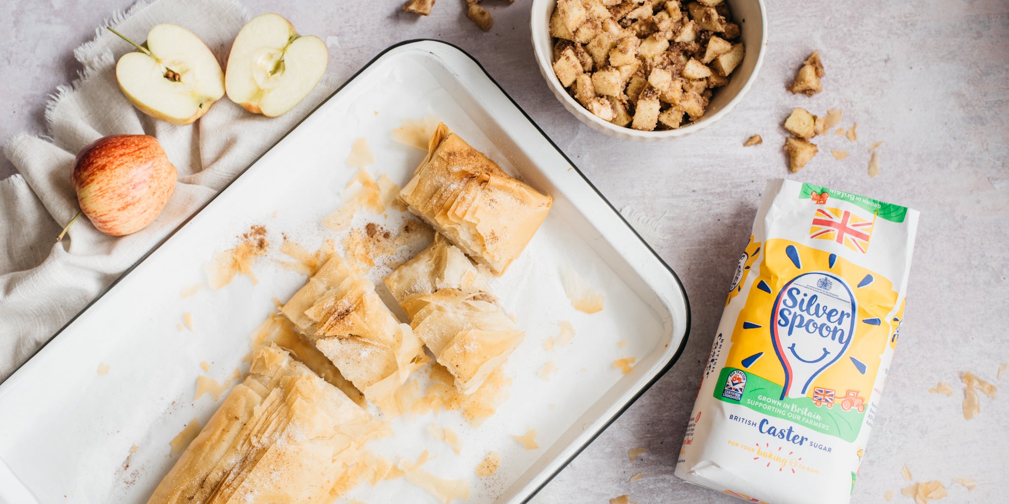 Apple strudel in baking tray surrounded by apples and pack of sugar