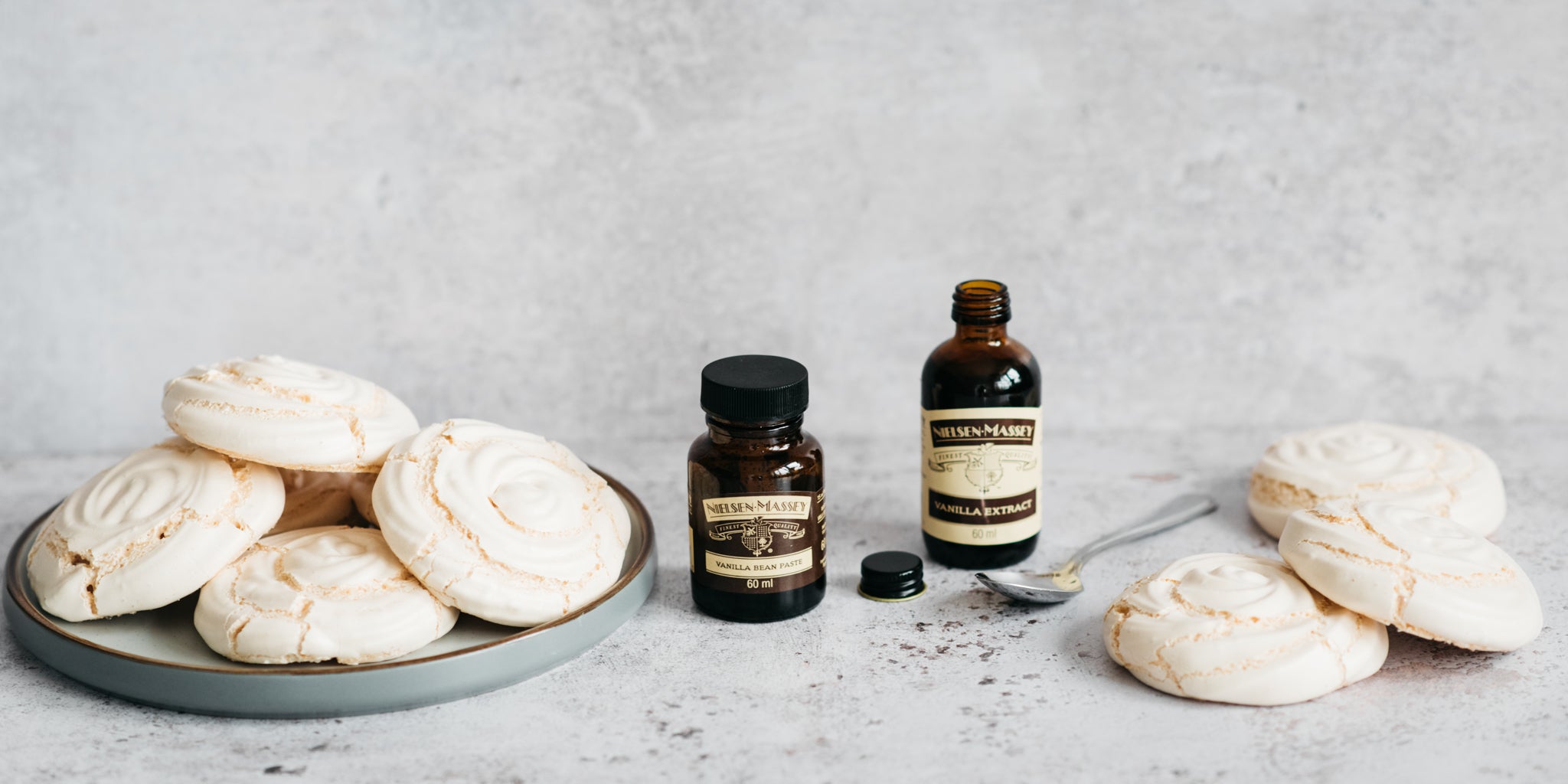 Plain baked meringue nests stacks on a plate to the left, vanilla extract bottles in the centre and 3 meringues stacked on the right