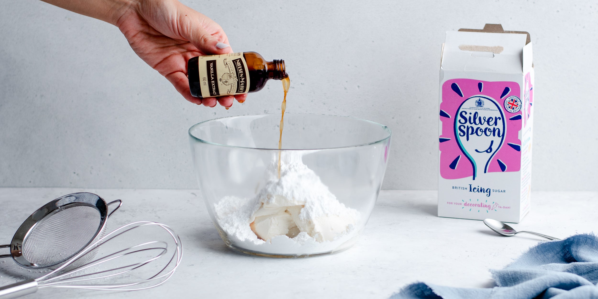 Vanilla extract pouring into a glass bowl beside an open pack of sugar and baking equipment