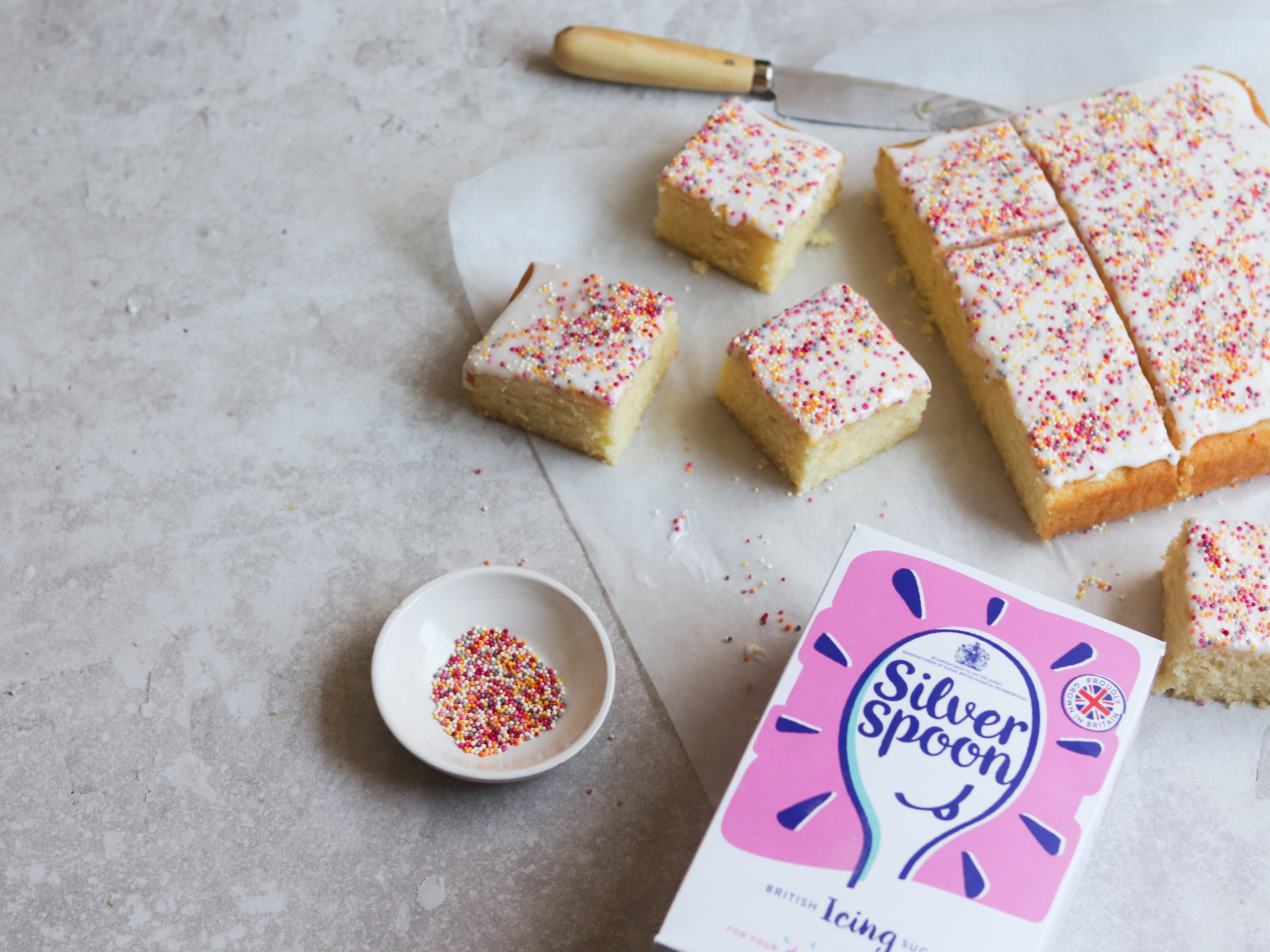 Sprinkle Cake from above view, on a sheet of baking paper, next to a box of Silver Spoon Icing Sugar and a bowl of sprinkles