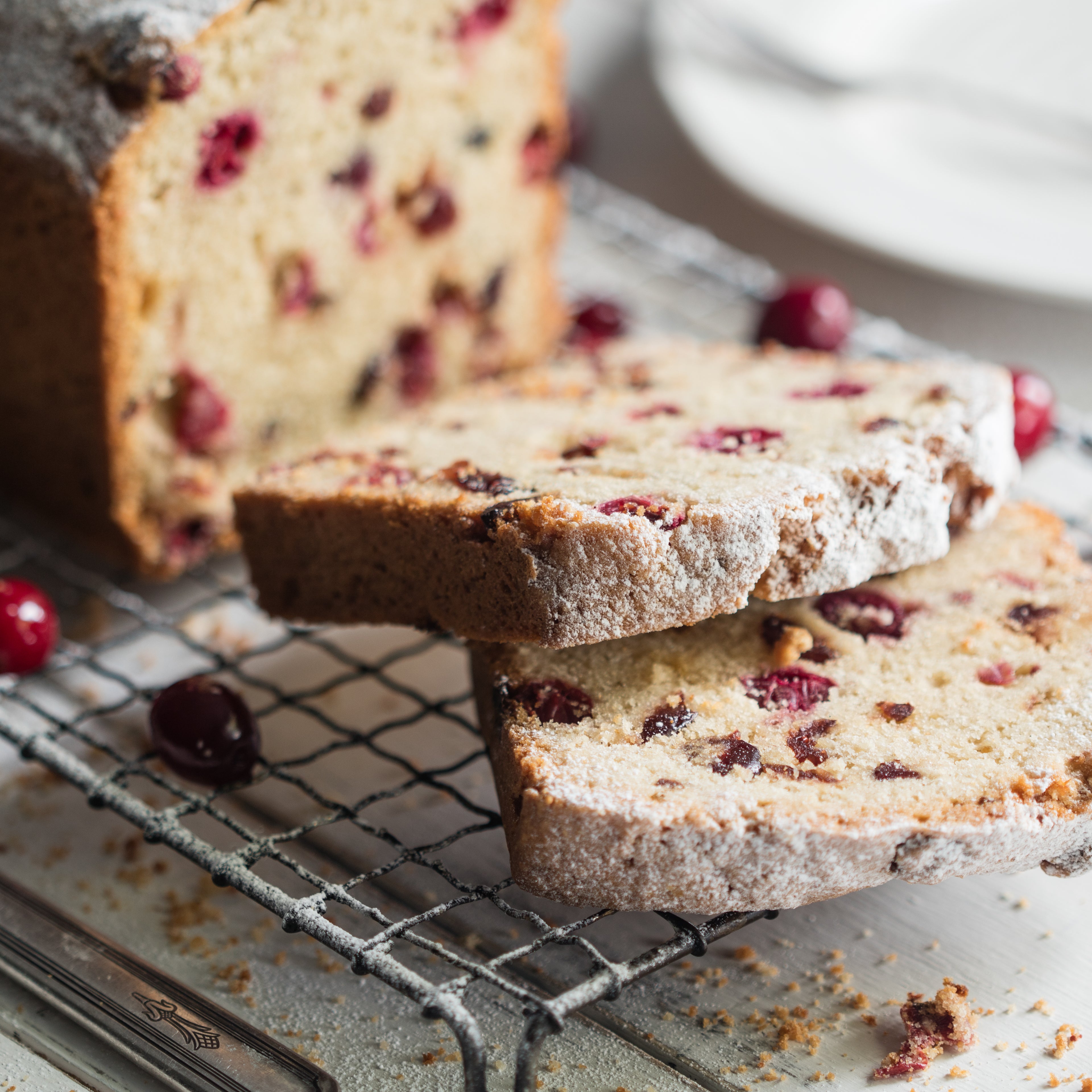 Two slices of cranberry loaf cake infront of the whole cake, dusted with icing sugar on a cooling rack