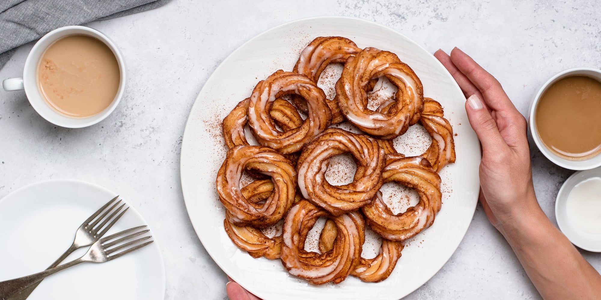 A plate of Apple Cider Crullers being held by hands. Dusted with cinnamon, next to cups of tea and cutlery