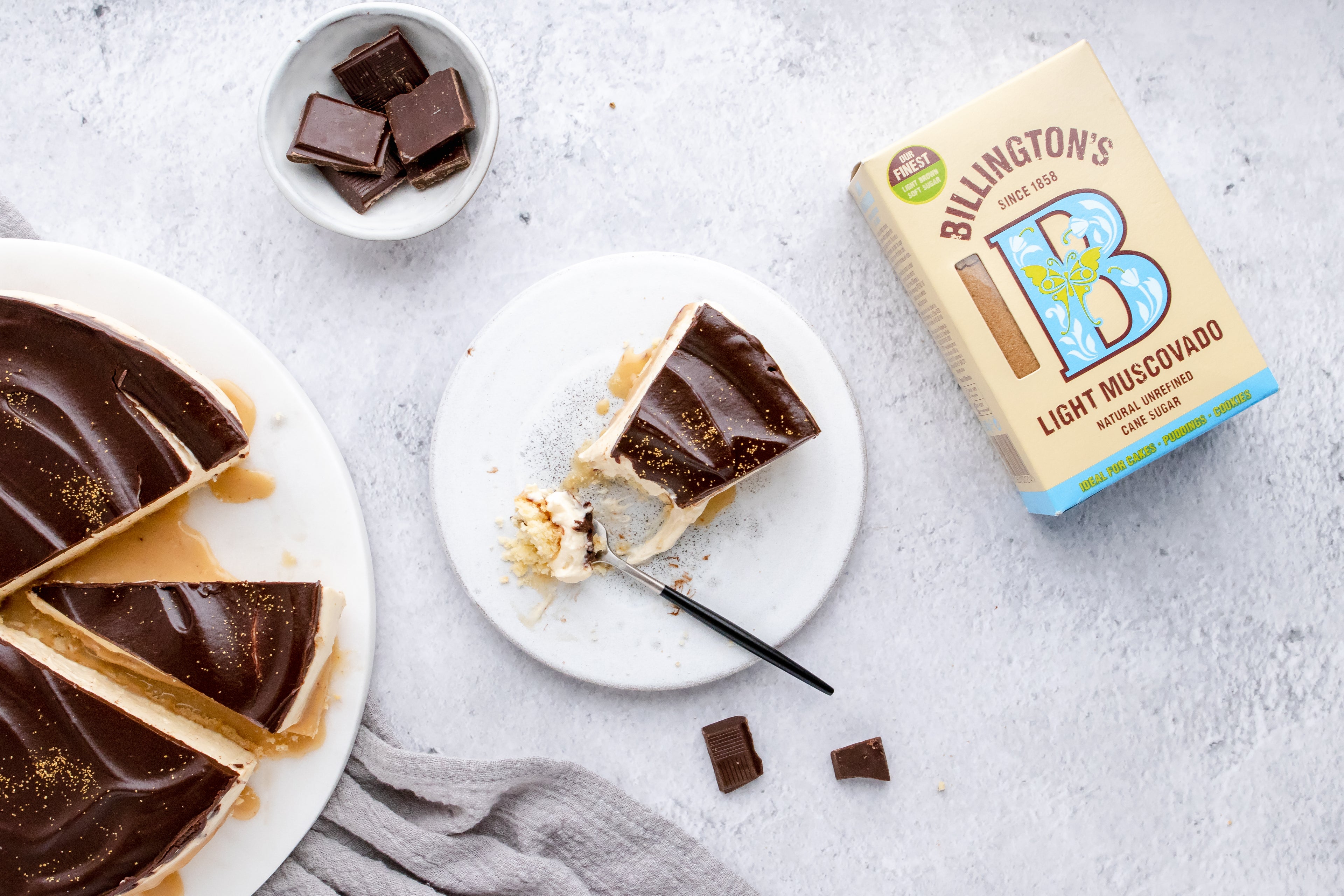 Millionaire's Cheesecake slice with a fork taking a piece out of it. Bowl of chocolate pieces and a box of Billington's Light Muscovado sugar