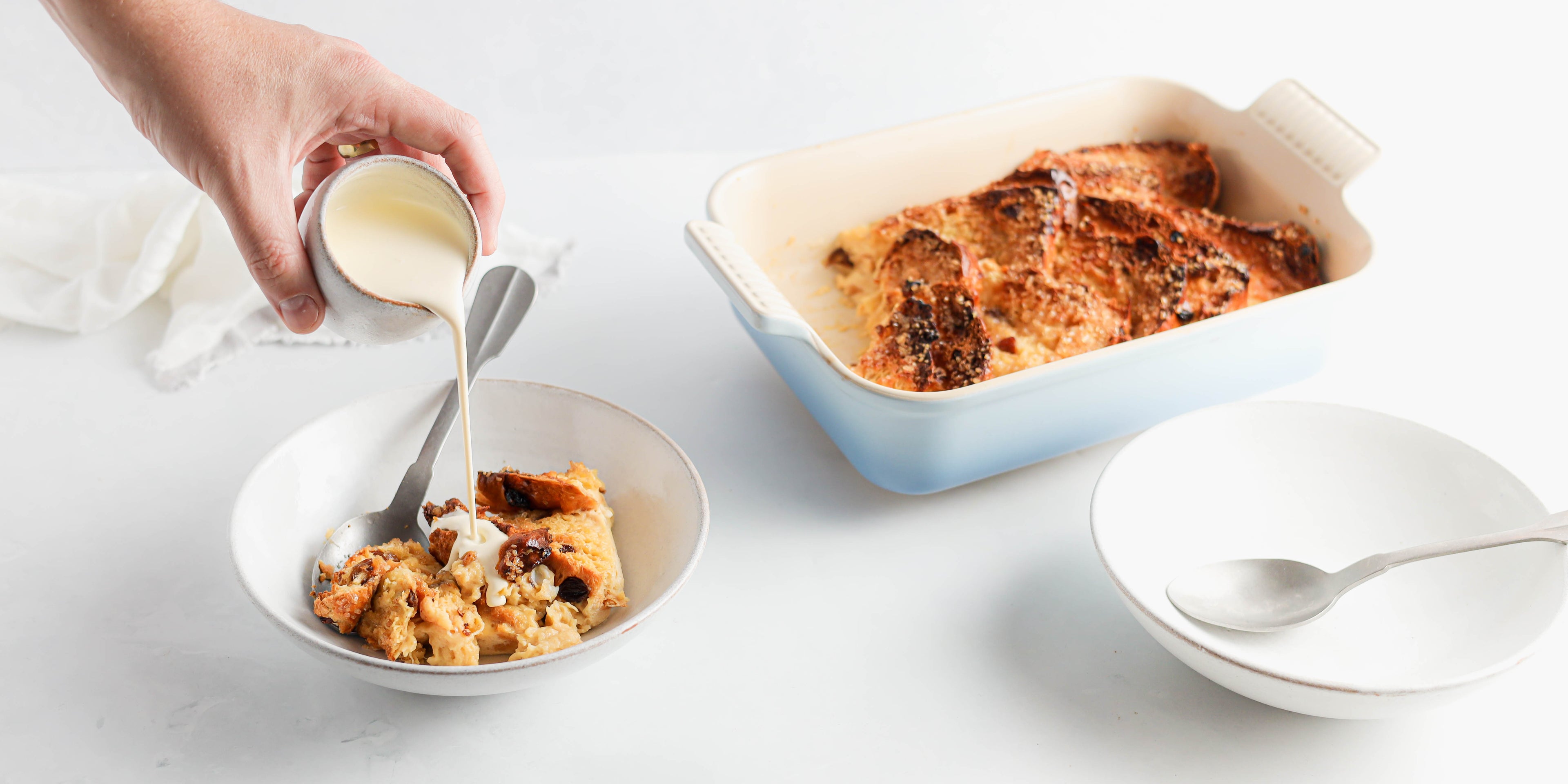 Panettone Bread & Butter Pudding in a bowl being poured with cream by a hand hold a cream jug