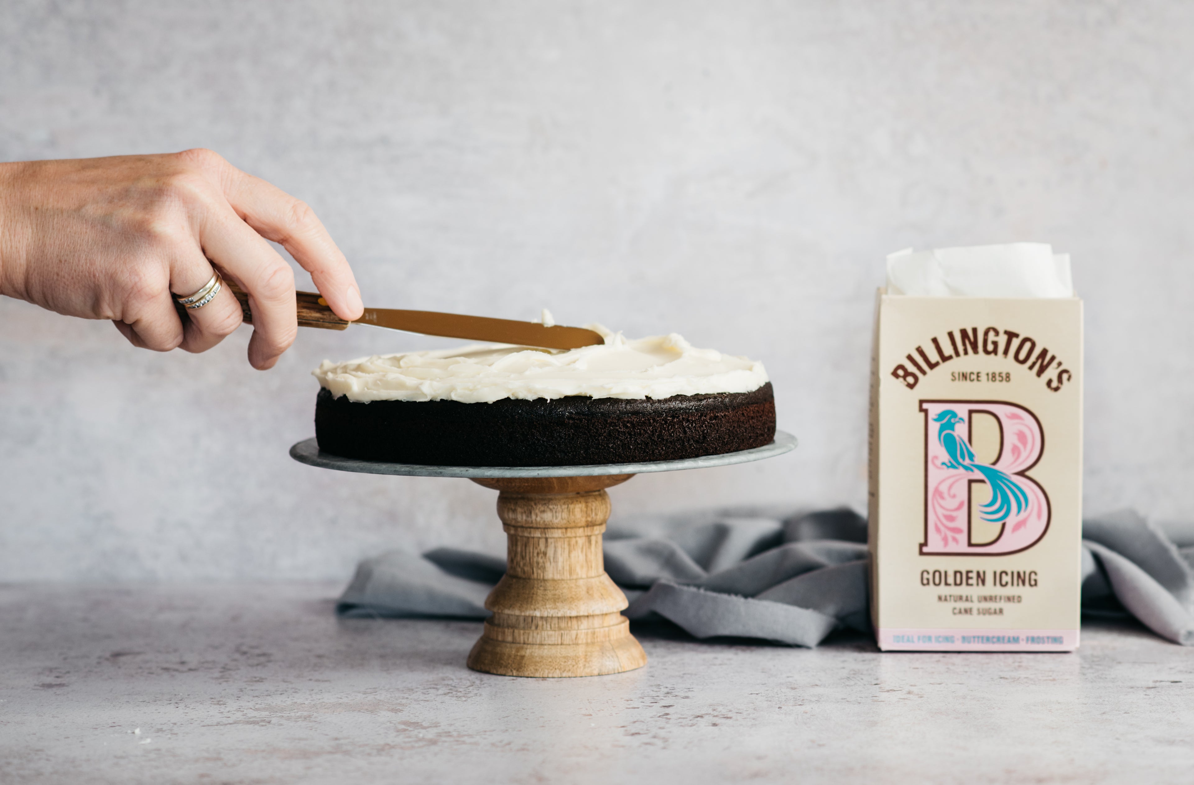 Hand using an offset spatula to smother a layer of Billington's Best Chocolate Cake with frosting using Billington's Golden Icing sugar