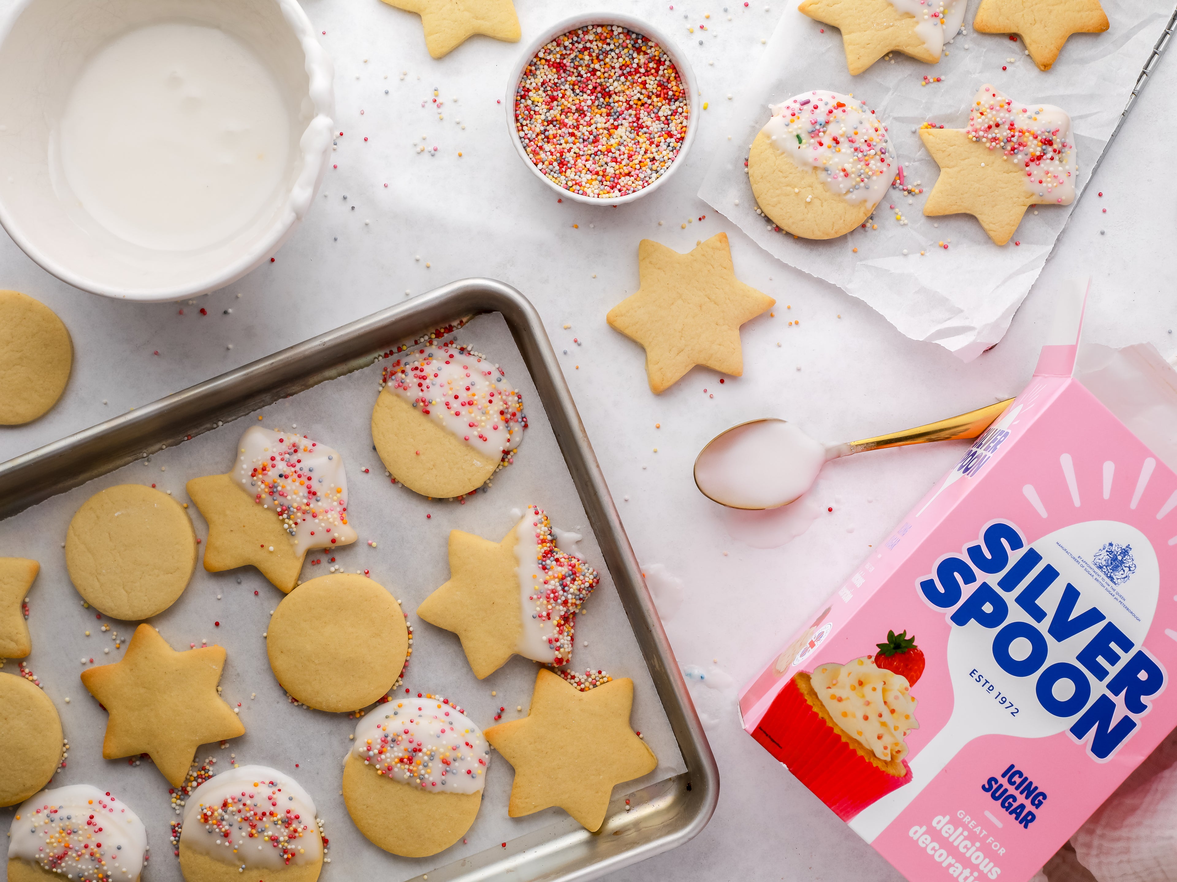 Biscuits decorated with icing and sprinkles on a baking tray beside icing sugar pack
