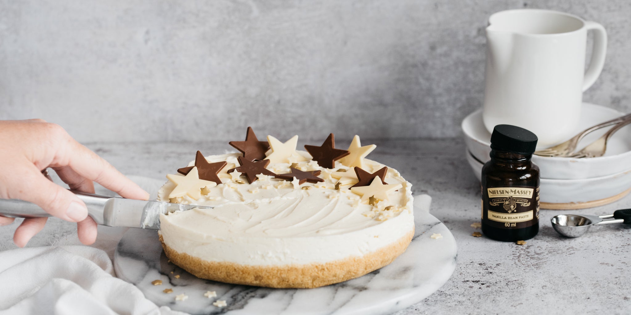 Hand with knife cutting into a vanilla cheesecake with chocolate star decorations. Bottle of vanilla bean paste in shot next to stack of crockery