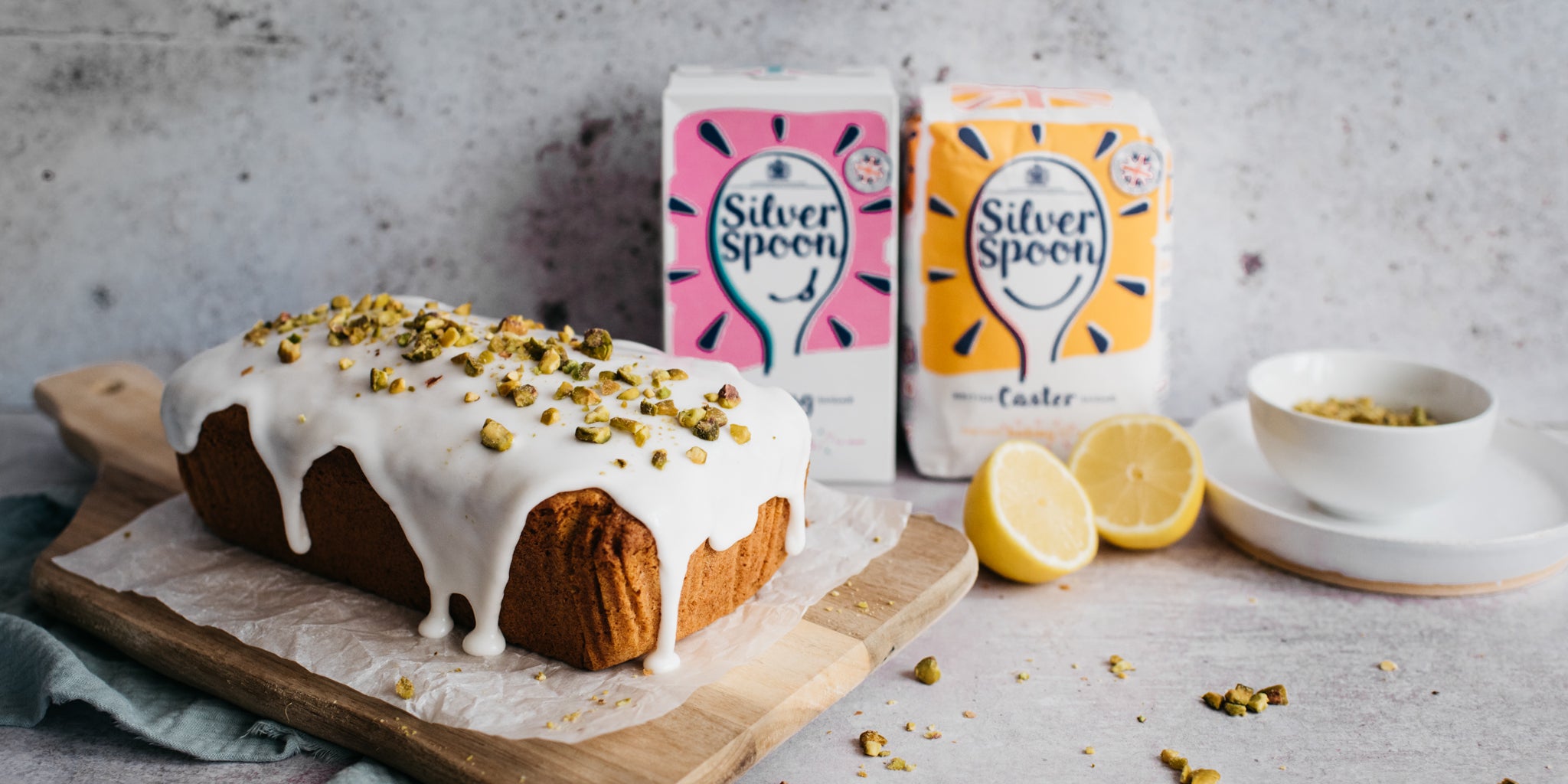 Gluten Free Vegan Lemon Drizzle Cake topped with nuts, next to a sliced lemon and a box of Silver Spoon Caster and Icing Sugar