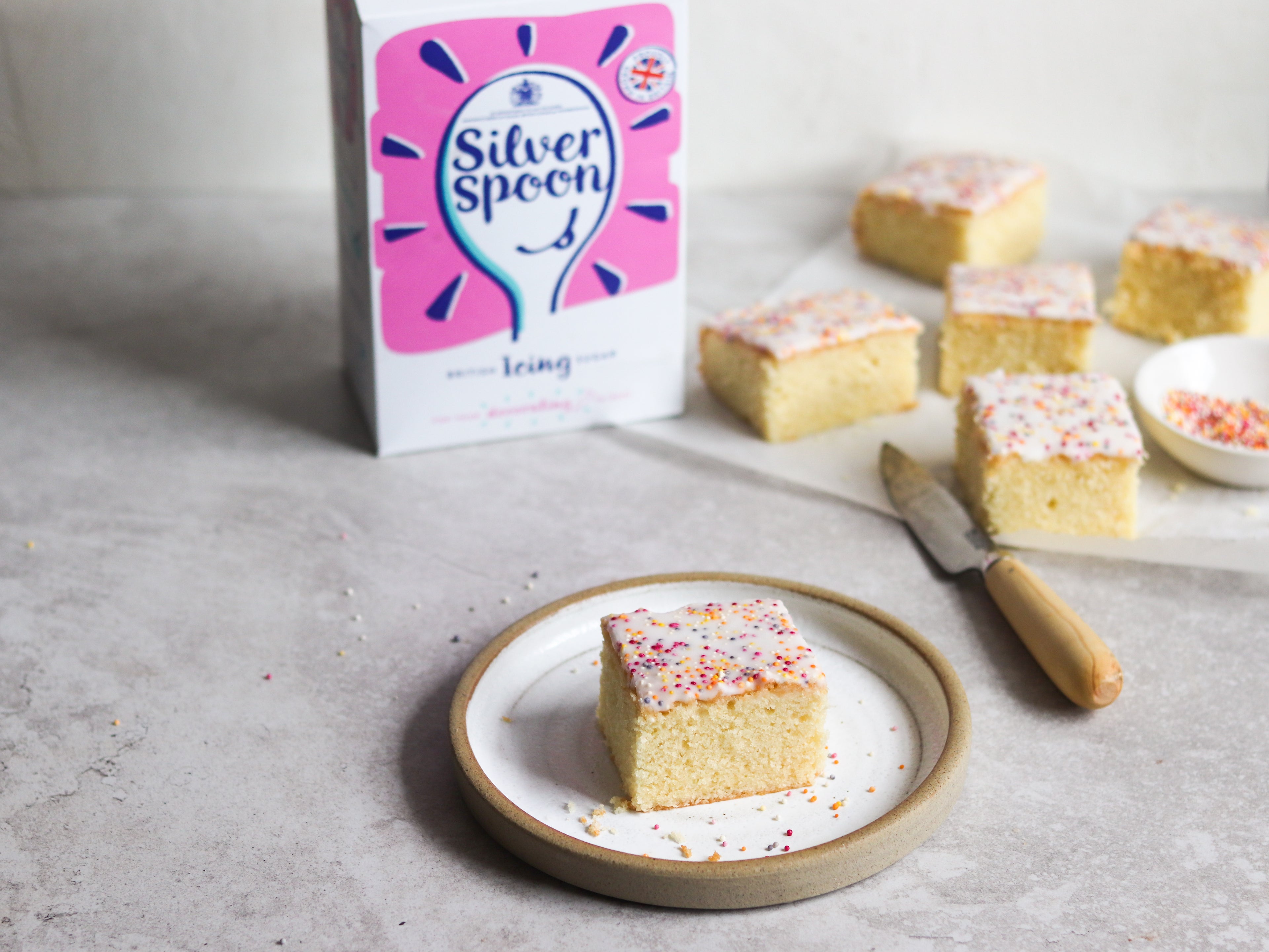 A Sprinkle Cake slice on a plate, with a box of Silver Spoon Icing Sugar in the background