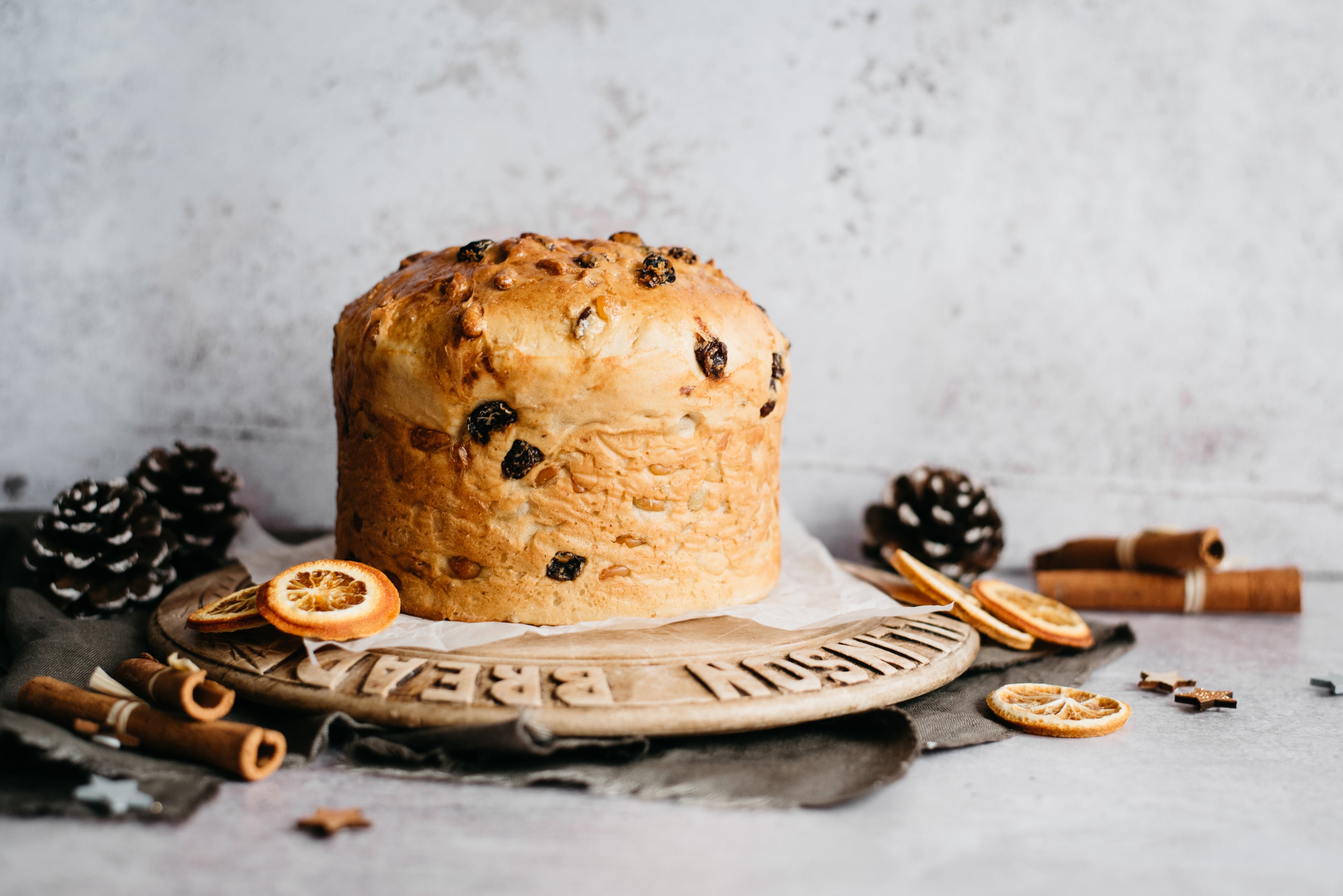 Close up of a golden baked Panettone filled with fruit, surrounded by festive pine cones and dried orange