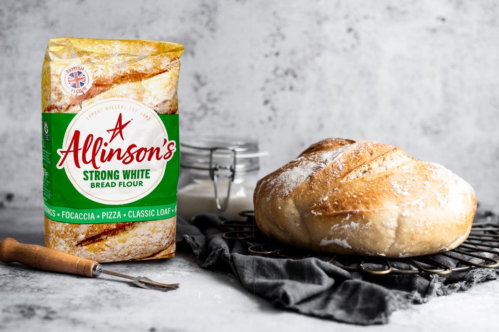 Bag of Allinson's Strong White Bread Flour next to a glass jar of sourdough starter and a sourdough loaf that sits on a charcoal grey piece of fabric.