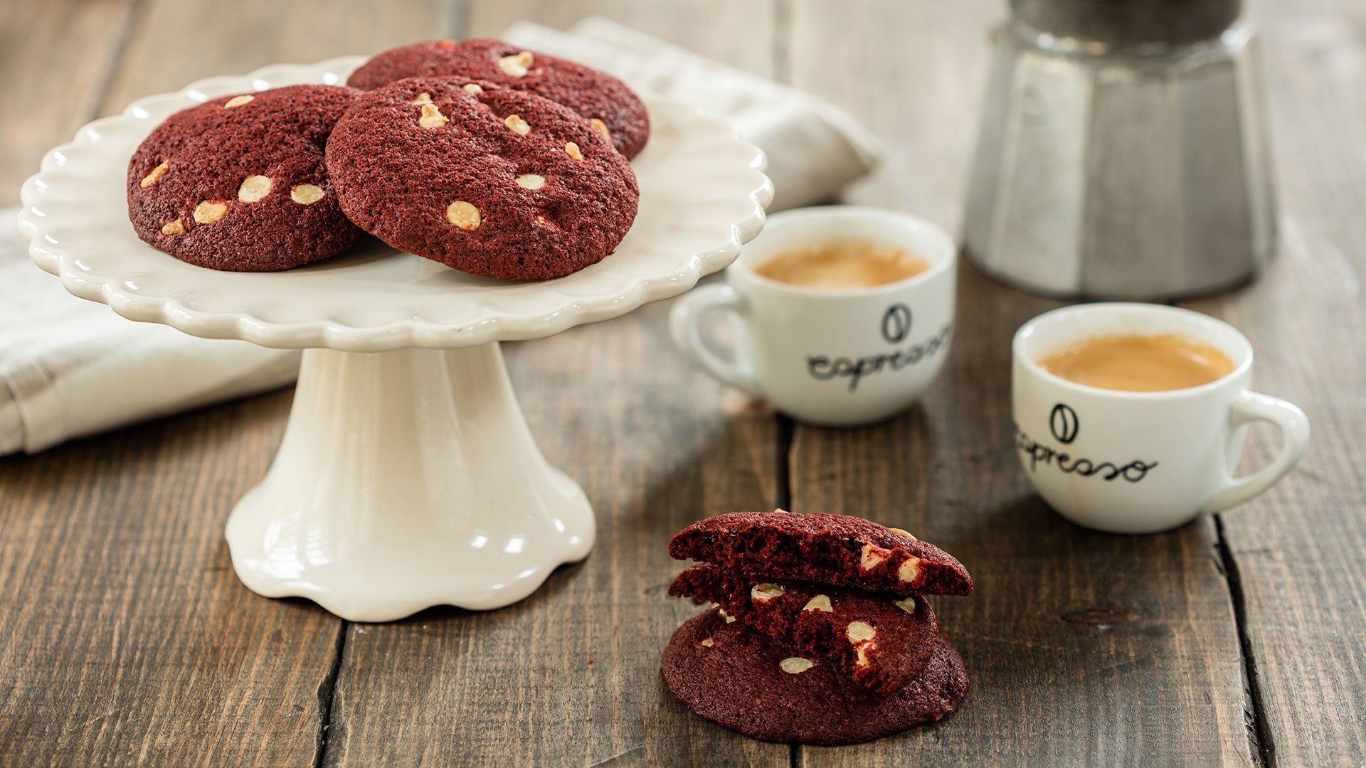 Red velvet cookies with white chocolate chips on a white cake stand next to two cups of coffee
