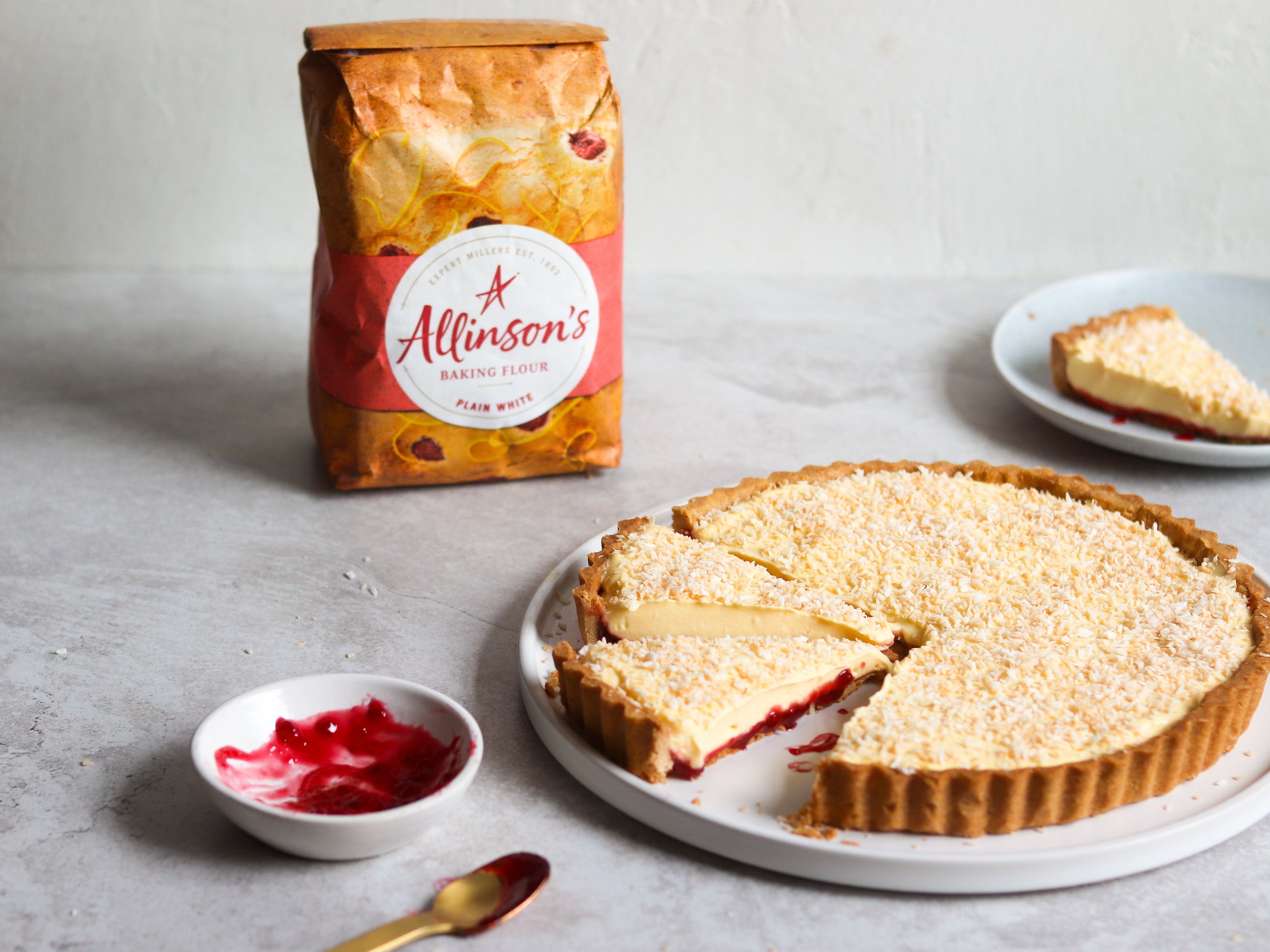 Manchester Tart with slice cut out of it, next to a bowl of jam and a spoon. Bag of Allinson's Plain Flour in background