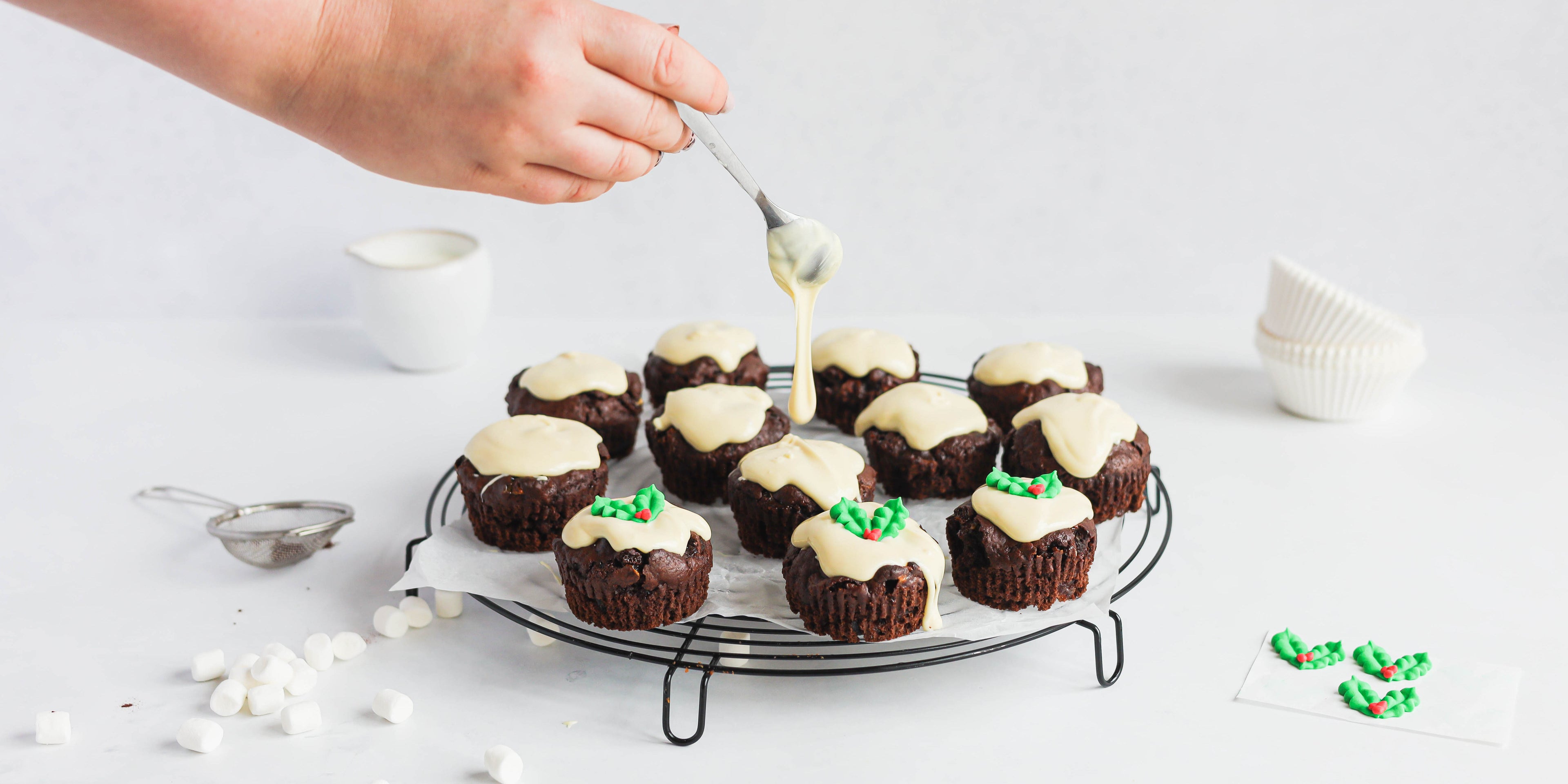 Mini Rocky Road Christmas Pudding Muffins being drizzled with a hand holding a spoon white chocolate