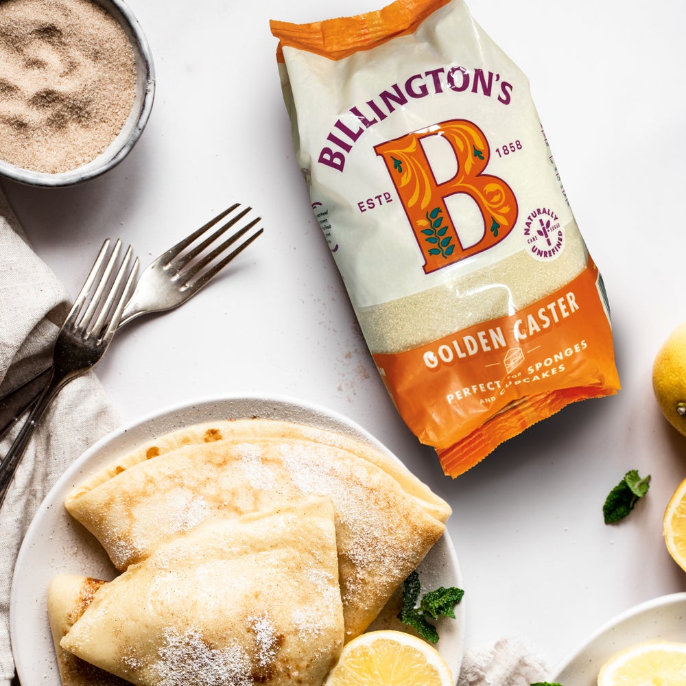 A bag of Billington's Golden Caster Sugar next to freshly made, folded pancakes covered in lemon and sugar shot from above, with some lemons to the side.
