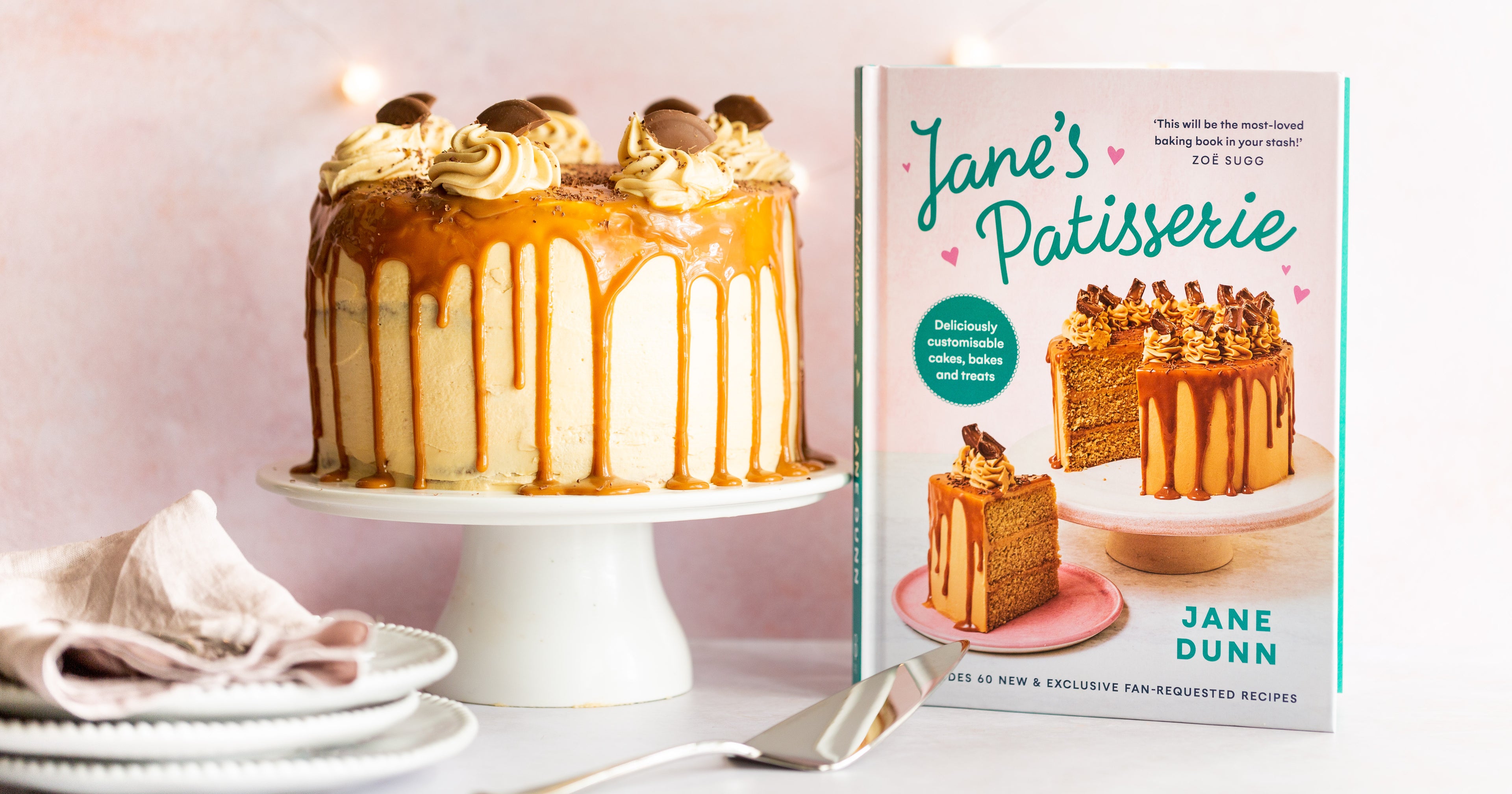 Cake on cake stand with caramel dripping down the side. A stack of white plates. A cake slice and a recipe book
