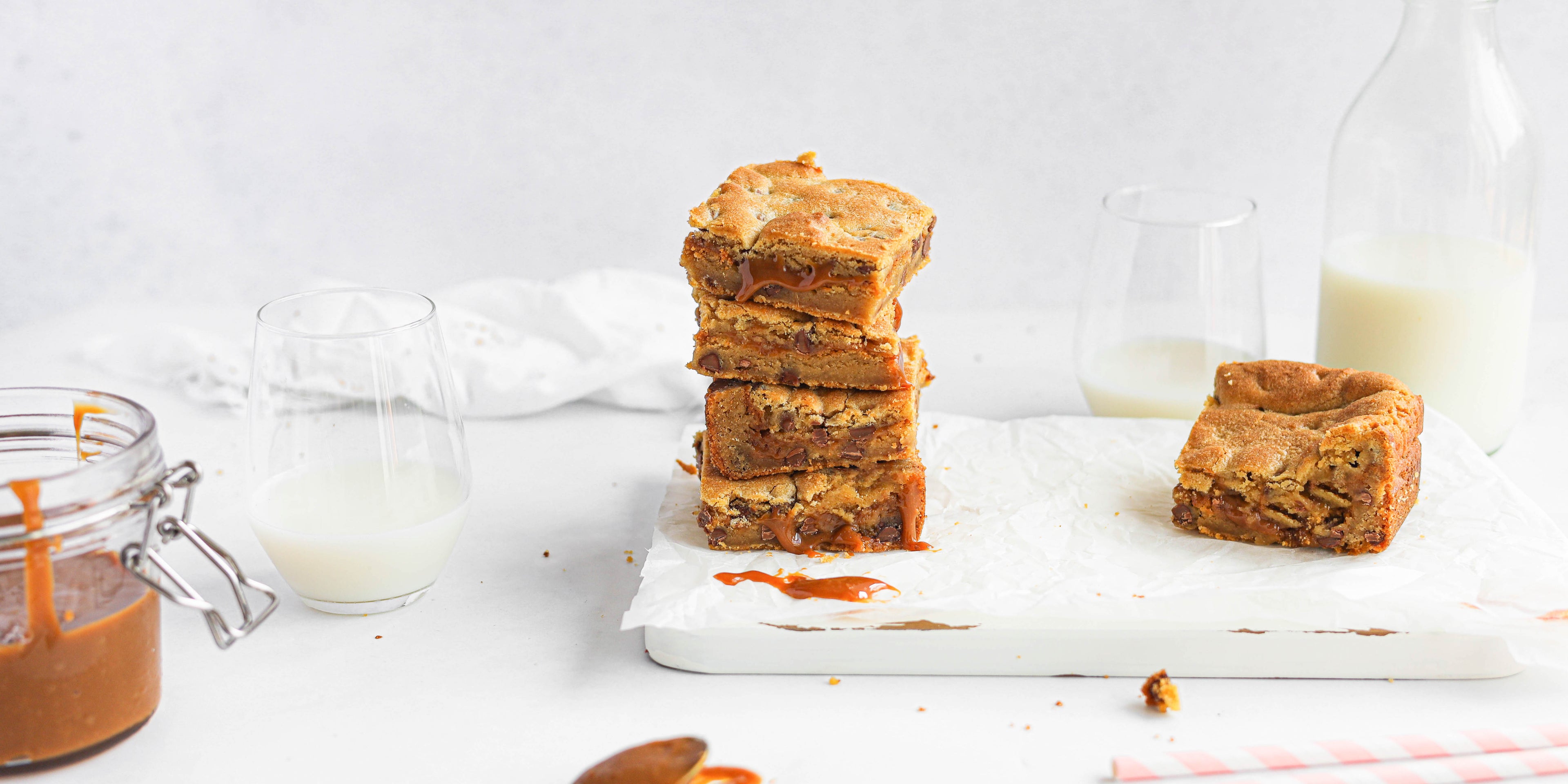 Stack of Salted Caramel Cookie Traybake on baking paper on a marble serving board, next to a jar of caramel and a half full glass of milk