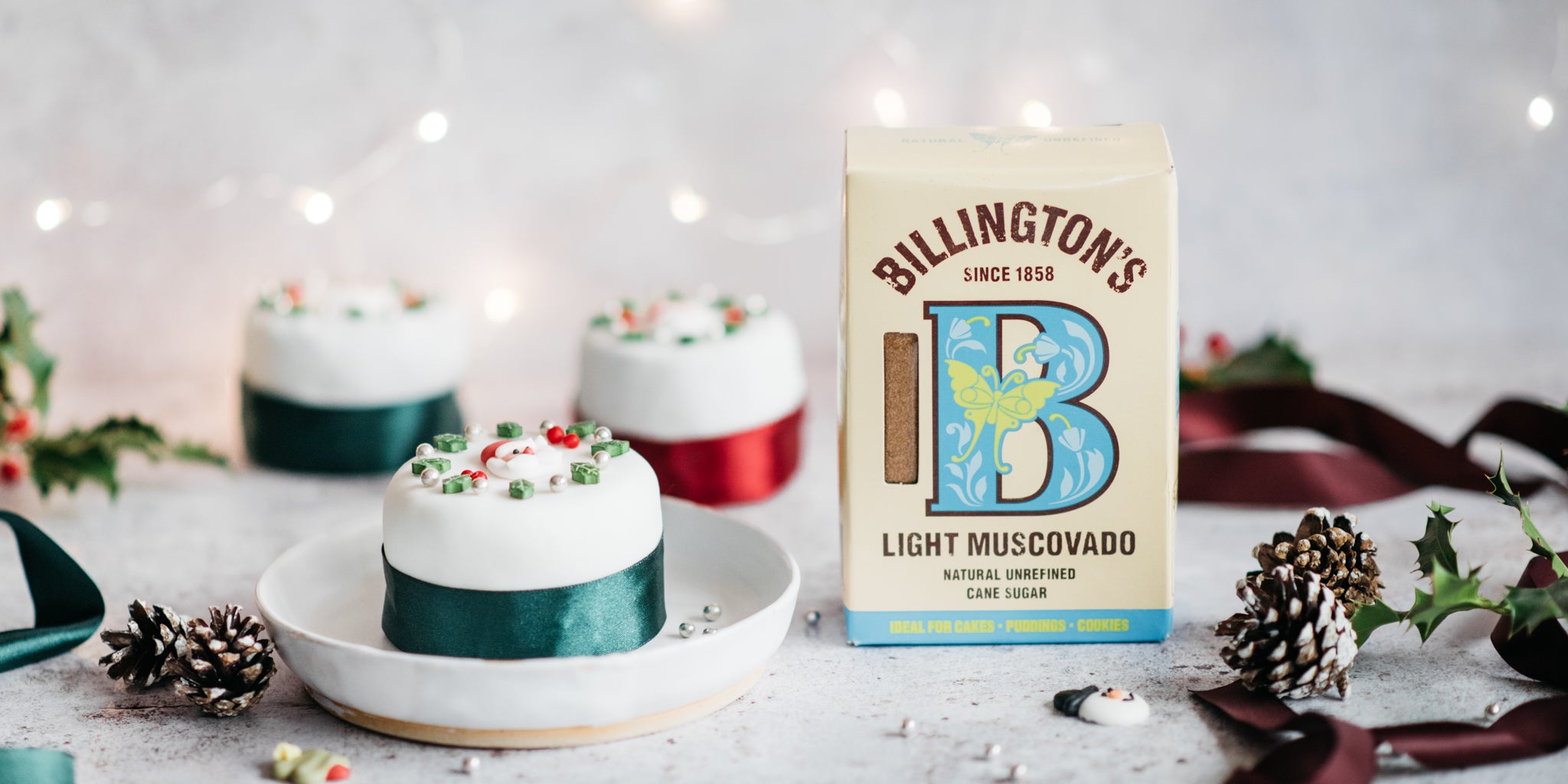 Mini christmas cake decorated with green ribbon on a white plate with a pack of light muscovado sugar beside it and festive decorations infront