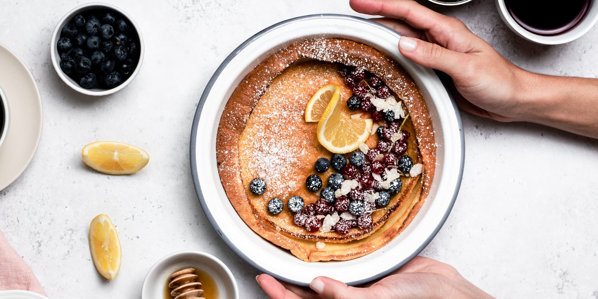 Hands holding plate of Dutch Baby Pancakes drizzled in honey, topped with berries and lemon, next to a cup of coffee, a honey drizzler and ramekin of blueberries