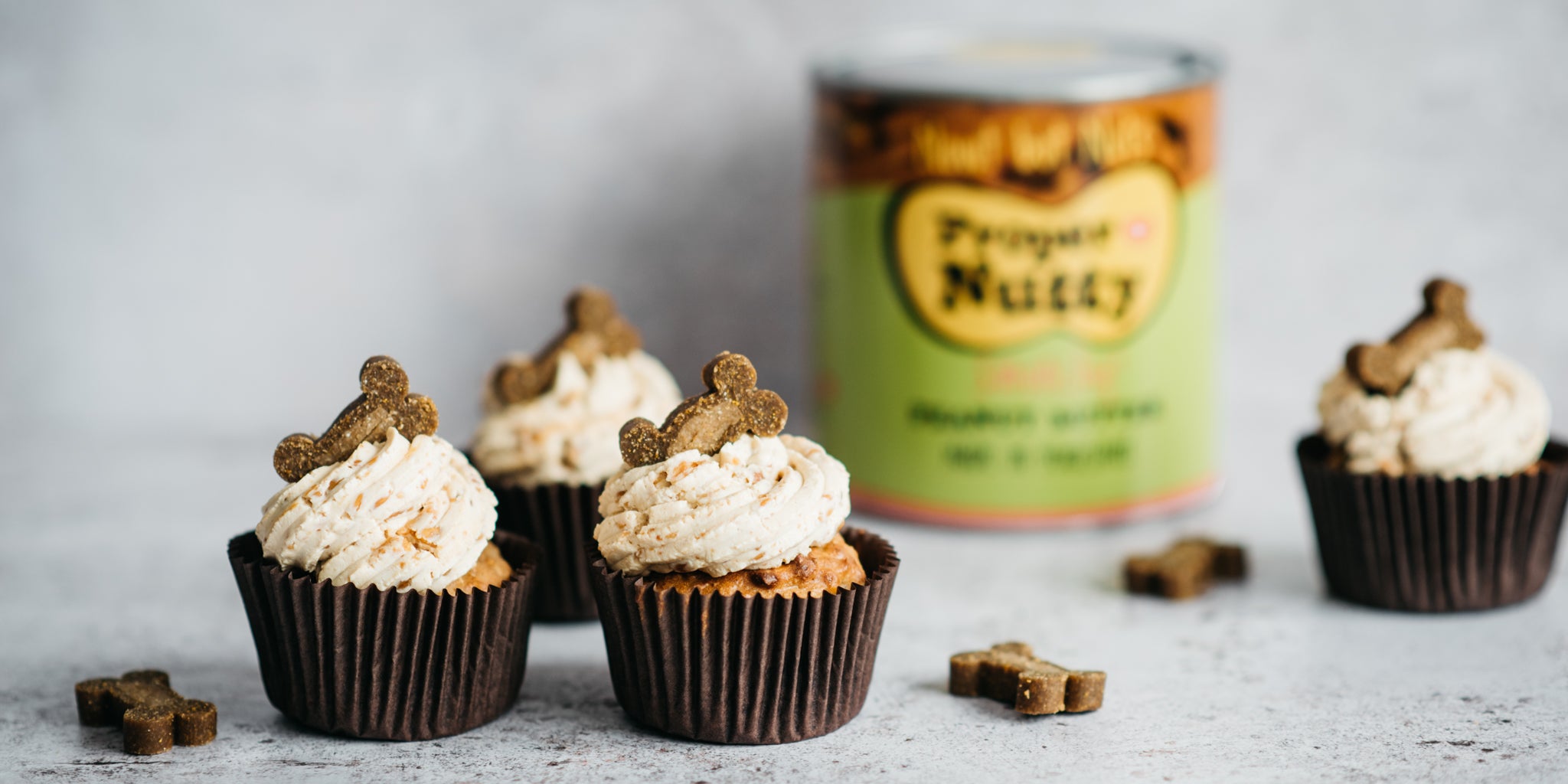 3 cupcakes with brown cases, frosting and dog treat topping in a group. Large tin of peanut butter in background next to another cupcake