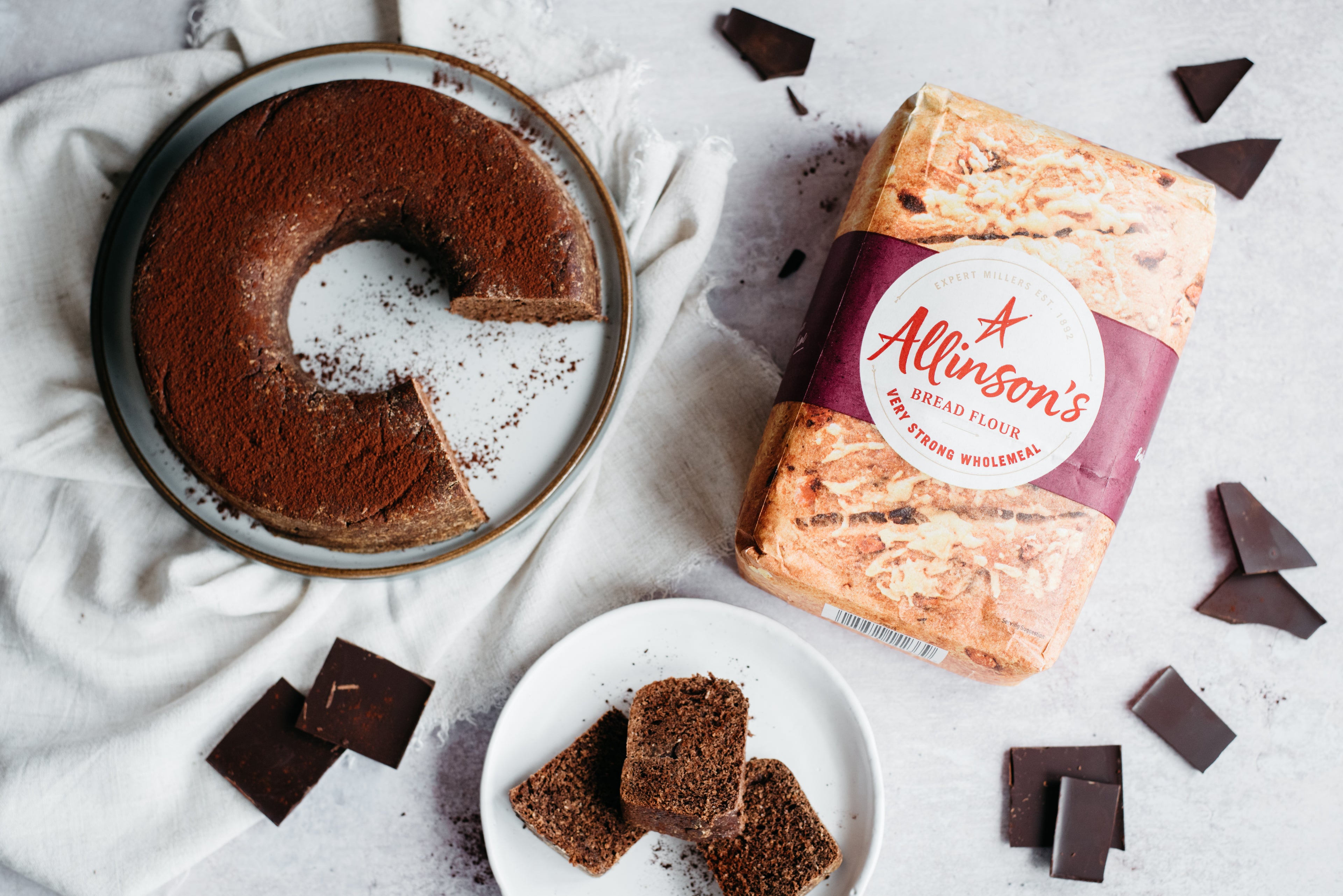 Top view of Chocolate Bread on a plate, with slices cut out and served on a plate, with dark chocolate pieces scattered around. Next to a bag of Allinson's very strong flour