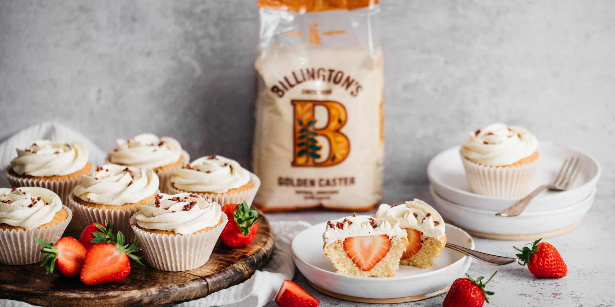 A circular plate of cupcake and strawberries. A bag of Billington's sugar in the background. A white plate with a cupcake cut in half and a fork on it. More plates in background with a cupcake on top