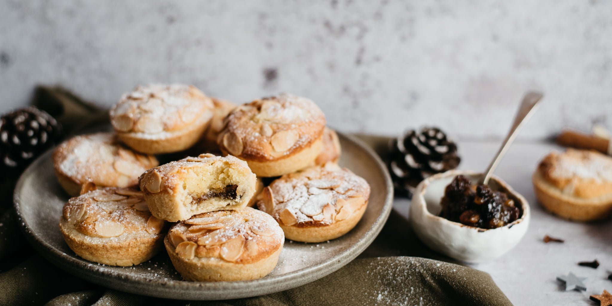 Close up of a batch of Billington's Frangipane Mince Pies by Benjamina dusted in icing sugar, next to a bowl of luxury mince meat