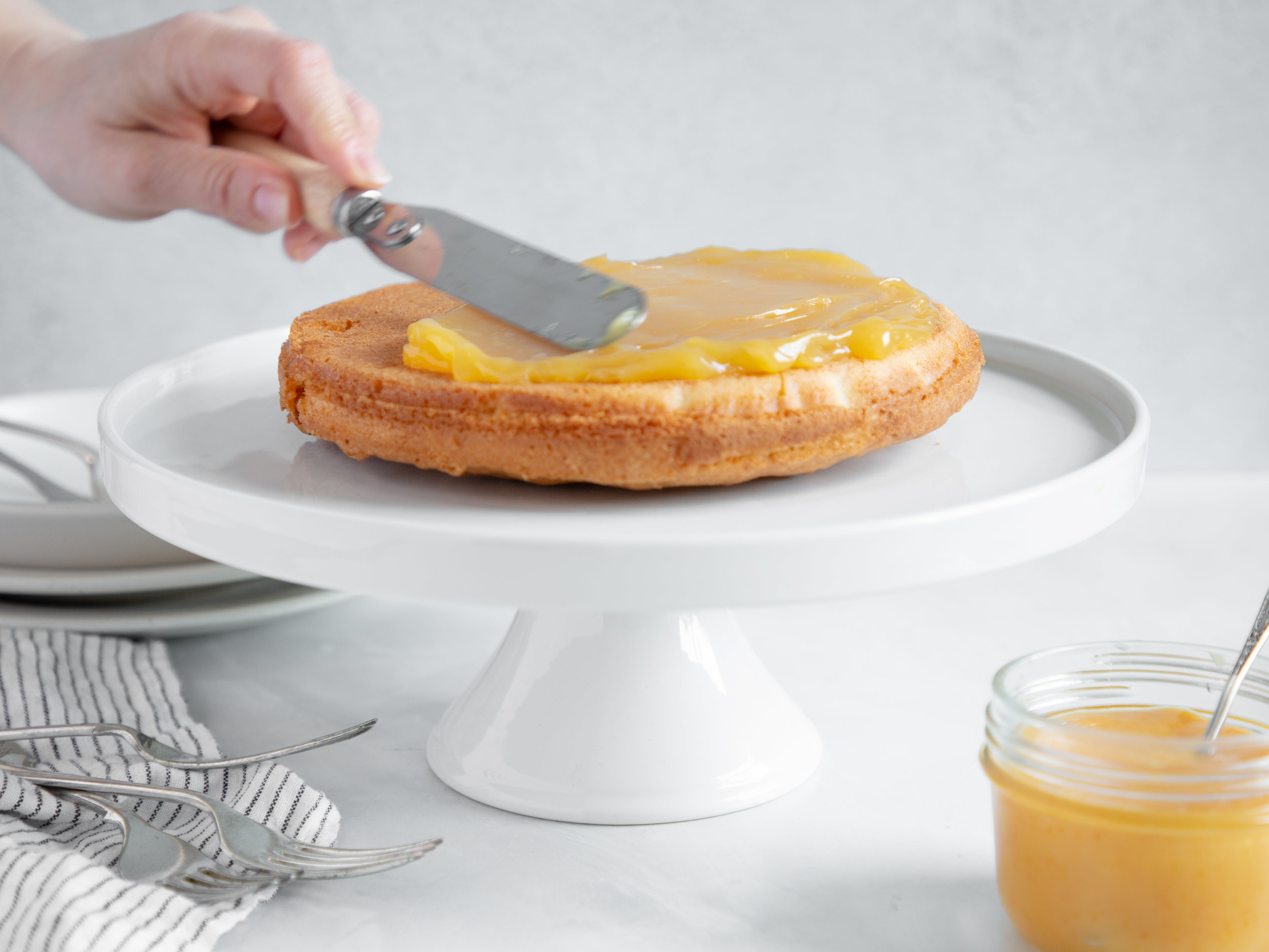 Classic Lemon Cake layer being smothered in lemon curd with a hand holding an offset spatula