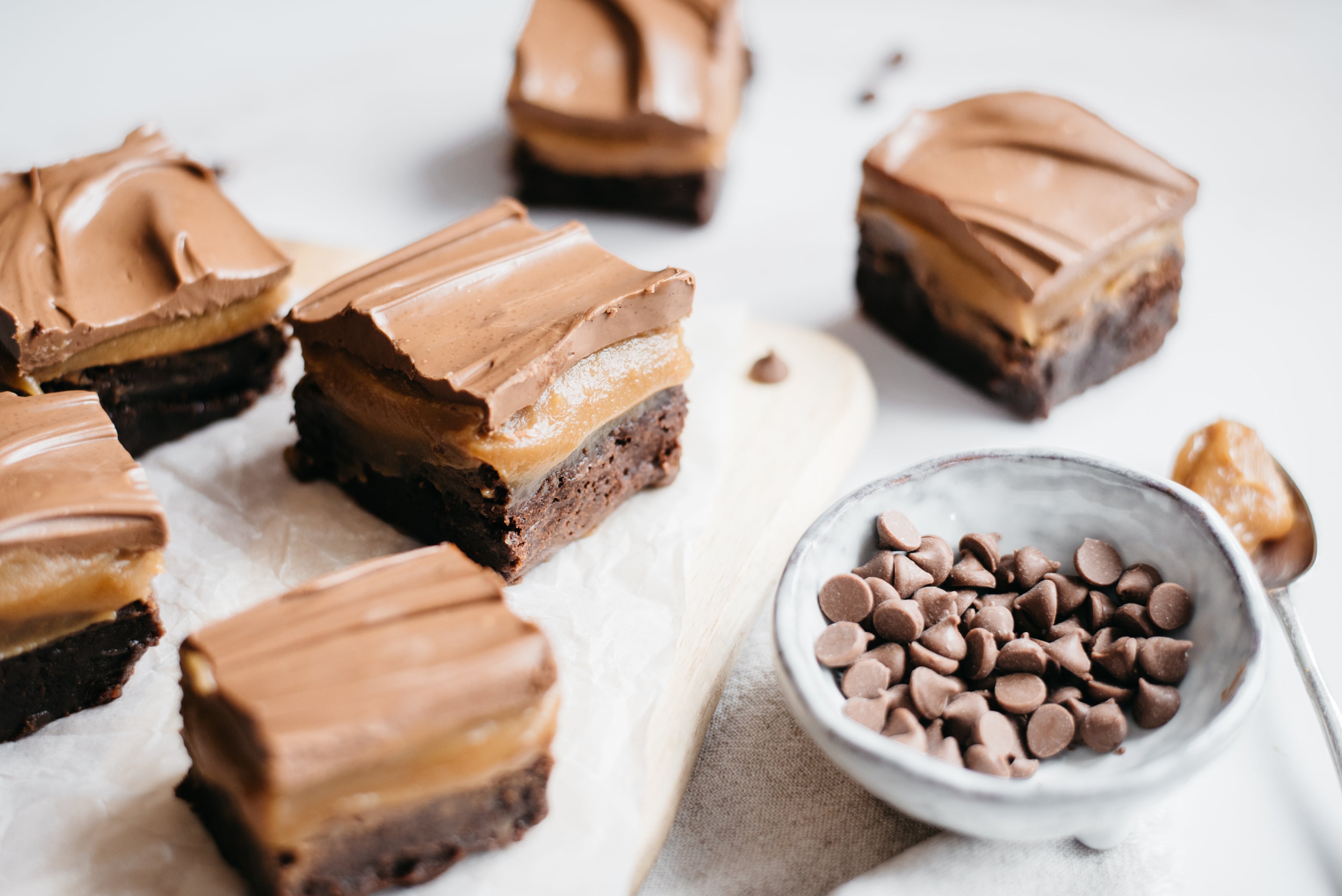 Slices of Brownie Based Millionaires Traybake with a bowl of chocolate chip, and a spoonful of caramel