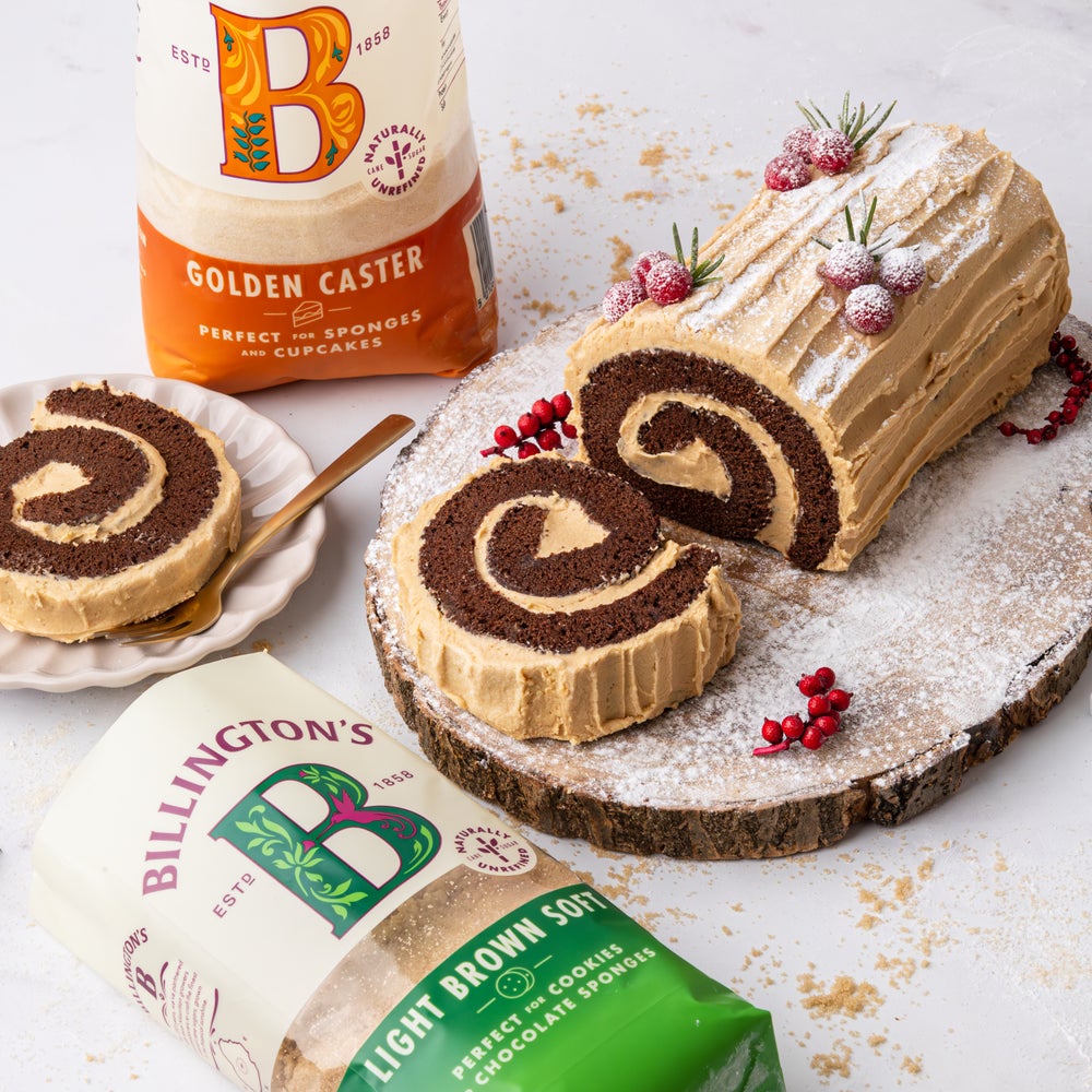 Biscoff Yule Log Cake sliced, with festive decorations on a wooden plinth next to two bags of Billington's sugars.