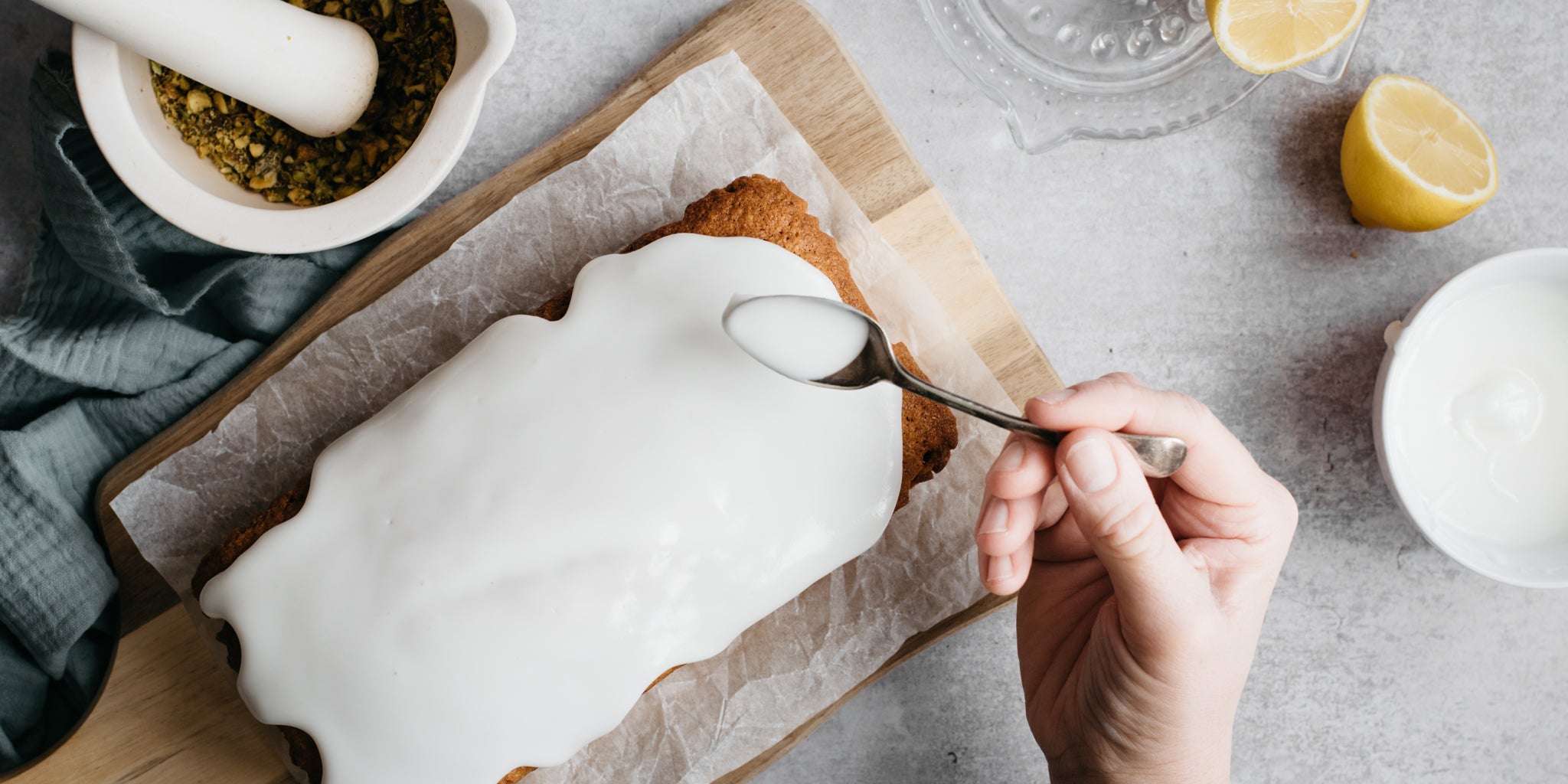 Hand drizzling a Gluten Free Vegan Lemon Drizzle Cake with Silver Spoon Icing Sugar, next to a sliced lemon and ground nuts